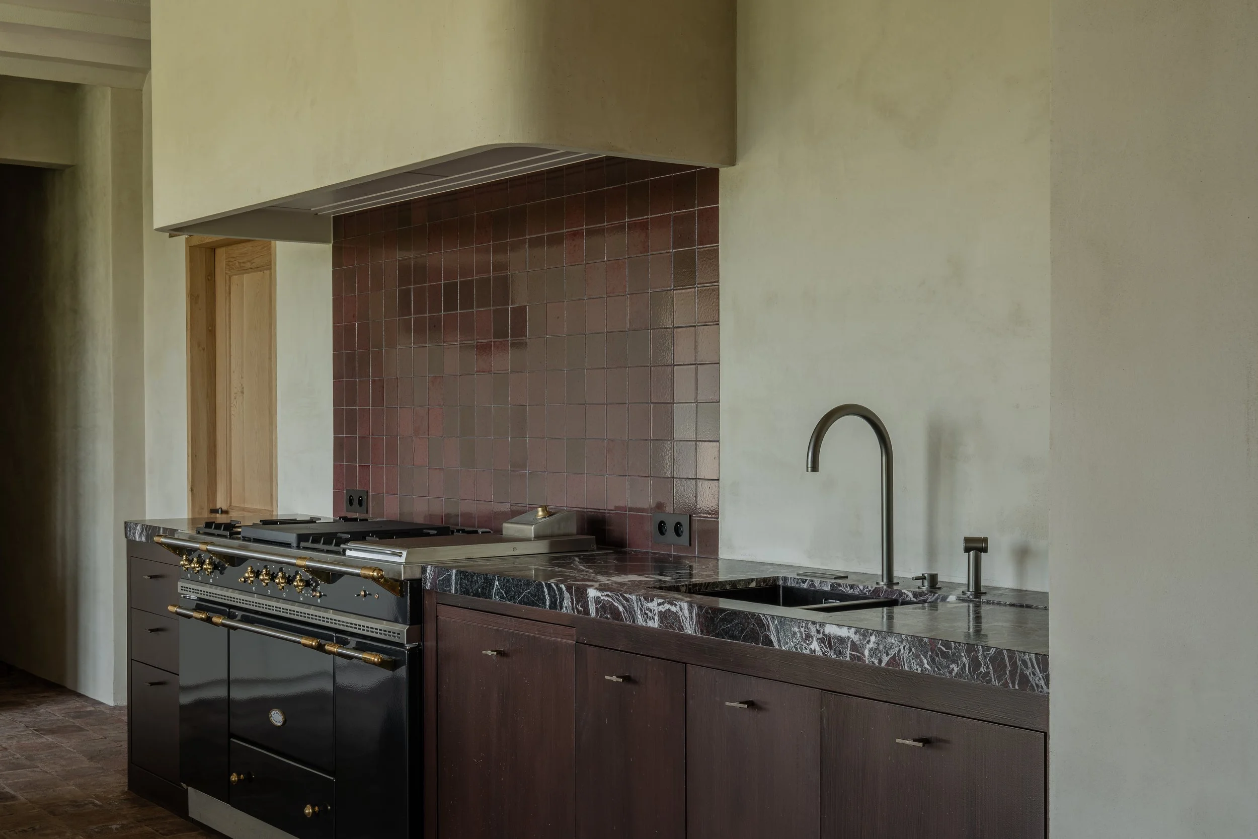 Kitchen with black stove, dark wood cabinets, marble countertop, red tiled backsplash, and beige walls.
