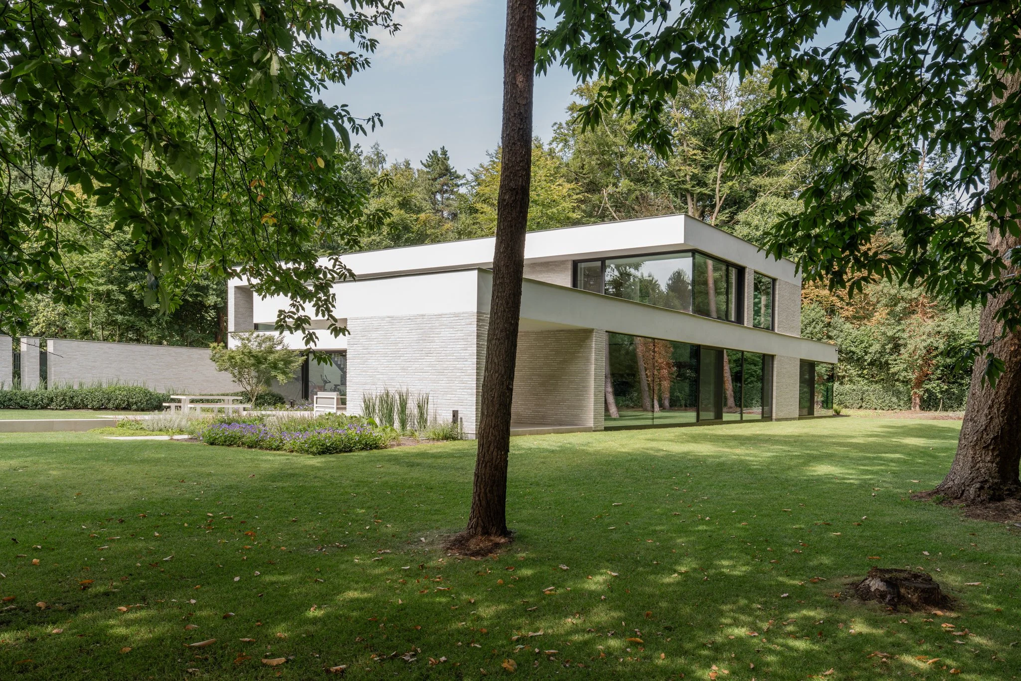 Modern white house with large glass windows, surrounded by greenery and trees, in a lush yard.