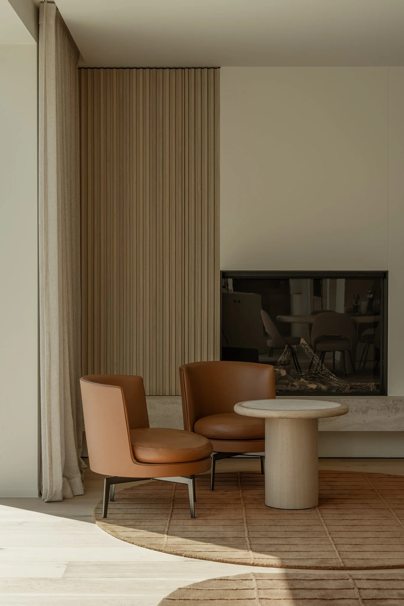 Interior corner with two brown leather chairs, a small round beige wooden table, a beige rug, beige curtains, and a modern fireplace.