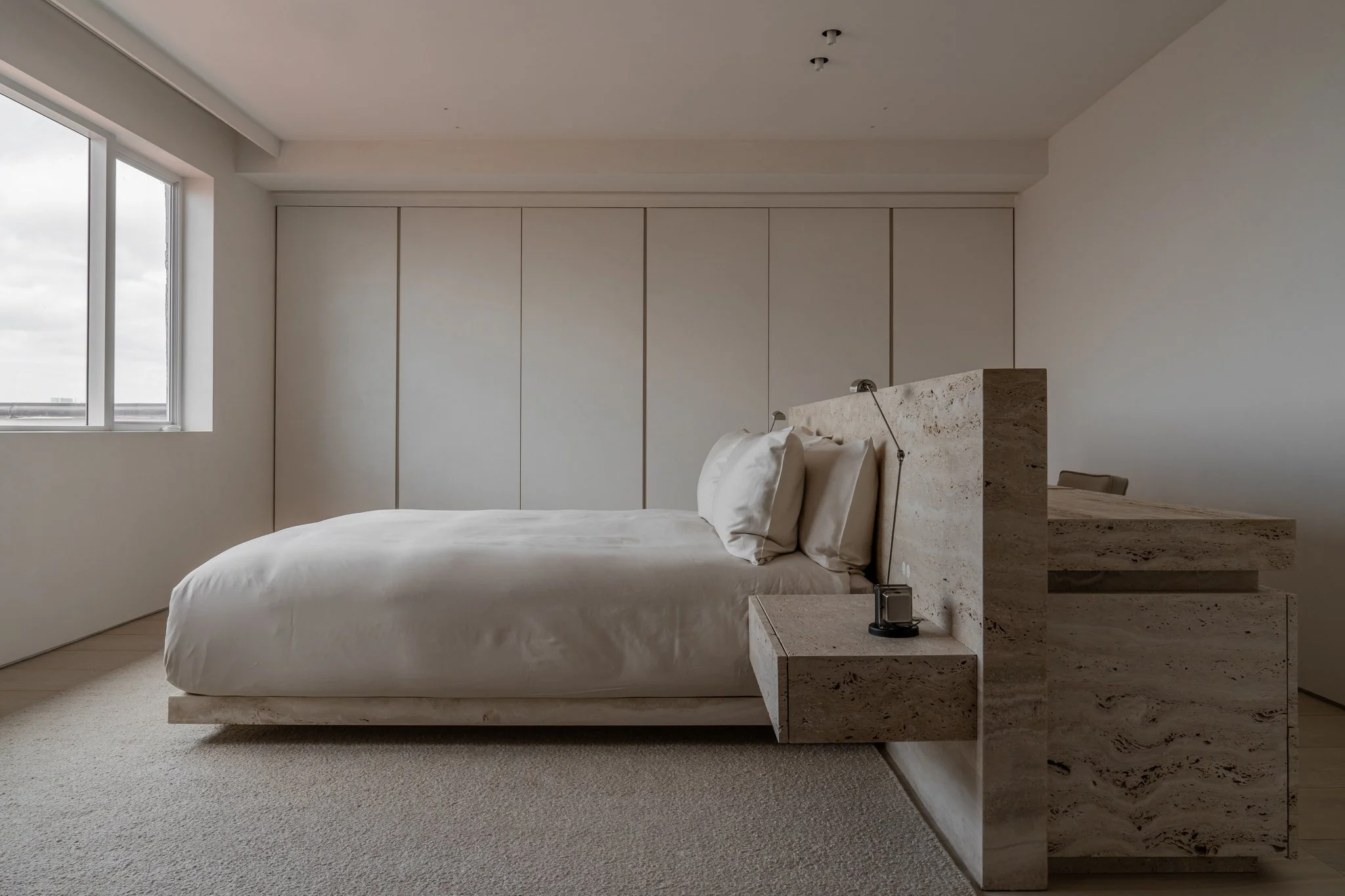 Minimalist bedroom with a large white bed, beige marble headboard, bedside table, and a window with city view.