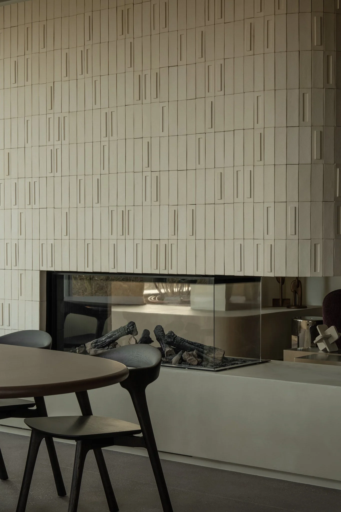 Modern fireplace with a glass enclosure and a decorative textured beige wall behind it, and a wooden dining table and chairs in the foreground.