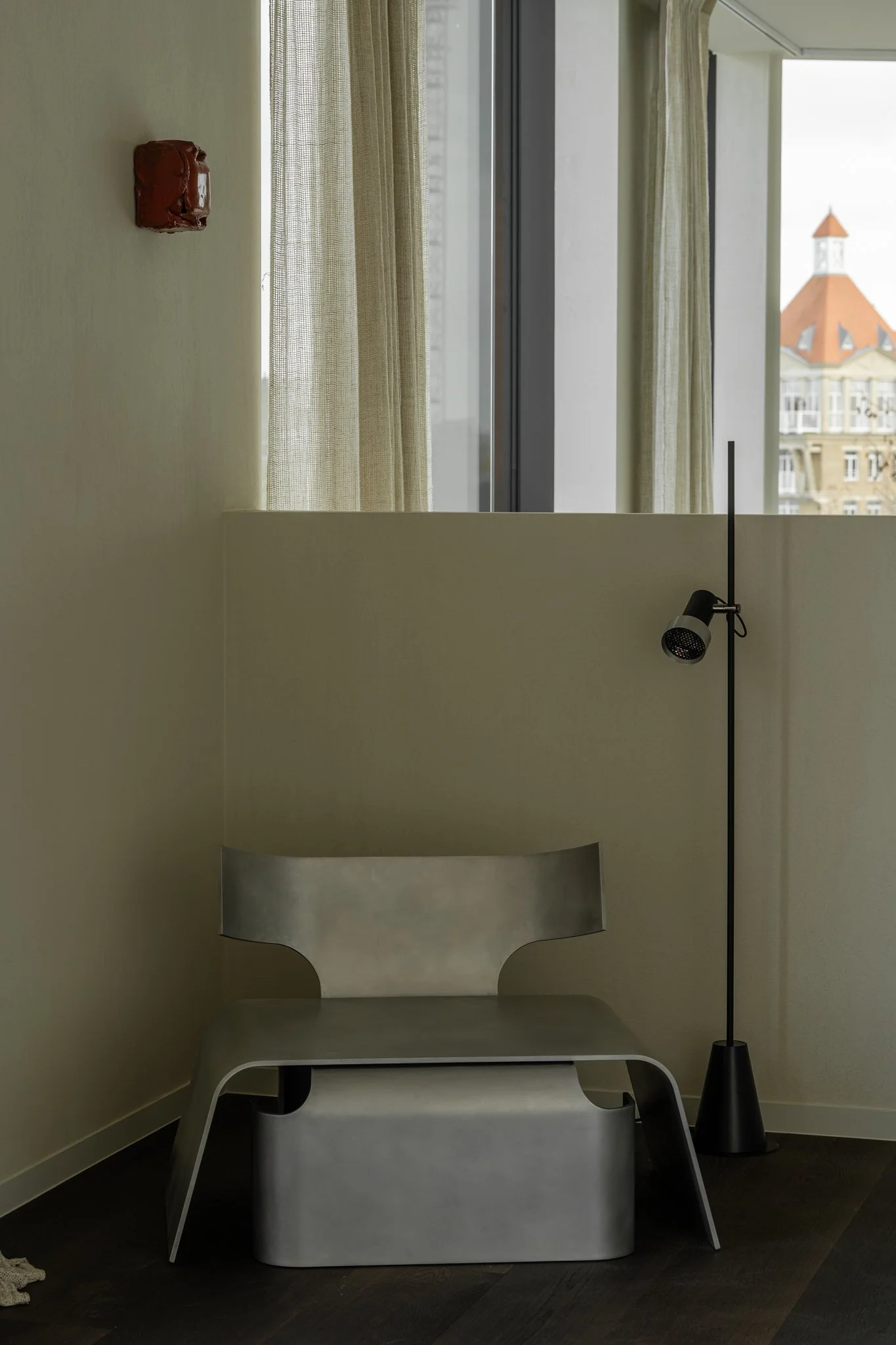 Modern interior corner with a metallic chair, a black floor lamp, beige curtains, and a window showing city buildings outside.