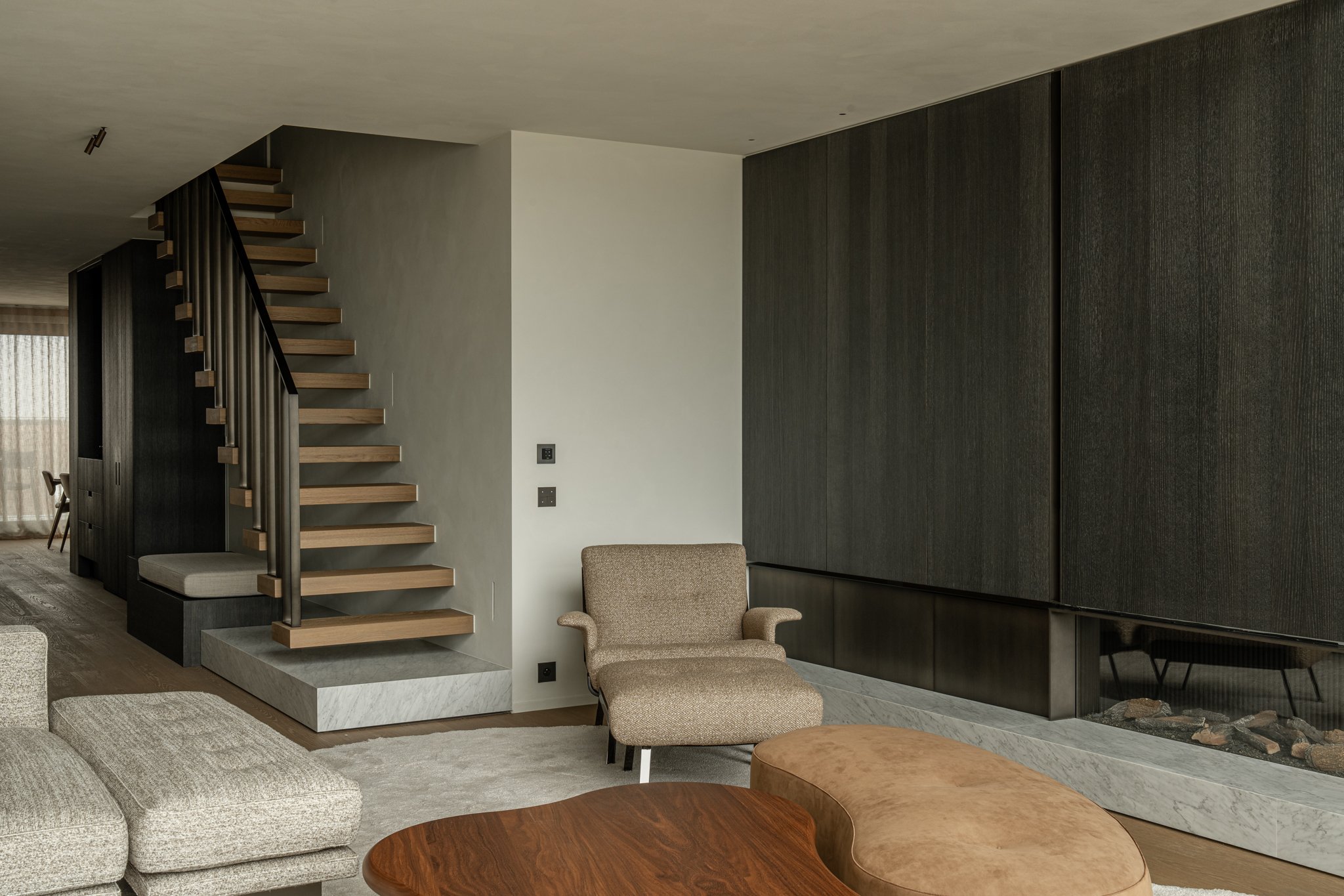 Living room with a staircase, beige armchair, wooden coffee table, and a modern fireplace with dark wood panels.
