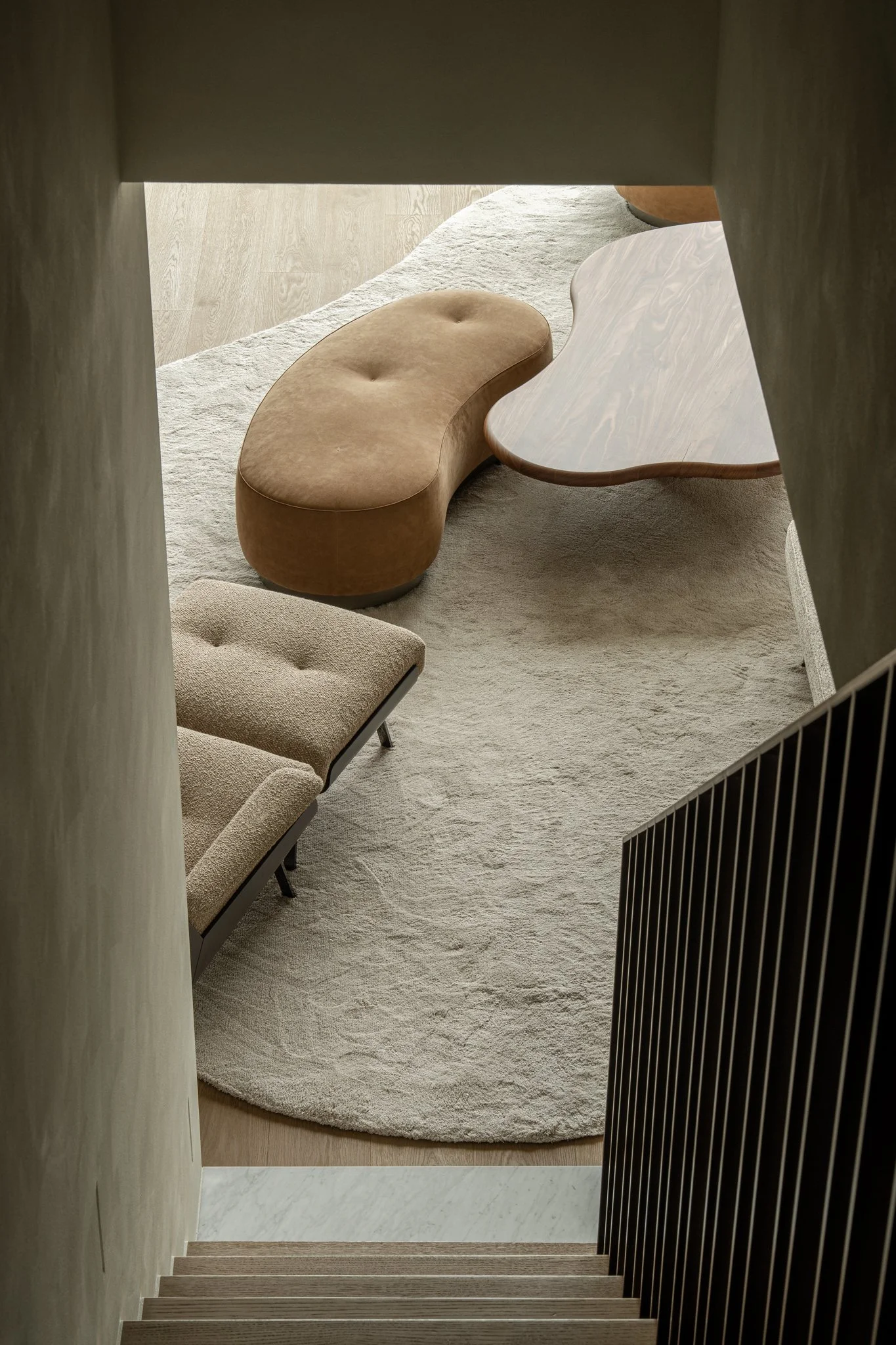 View of a modern living room from the staircase, featuring a beige oval bench, a matching two-seater sofa, a wooden coffee table, and a light-colored rug.