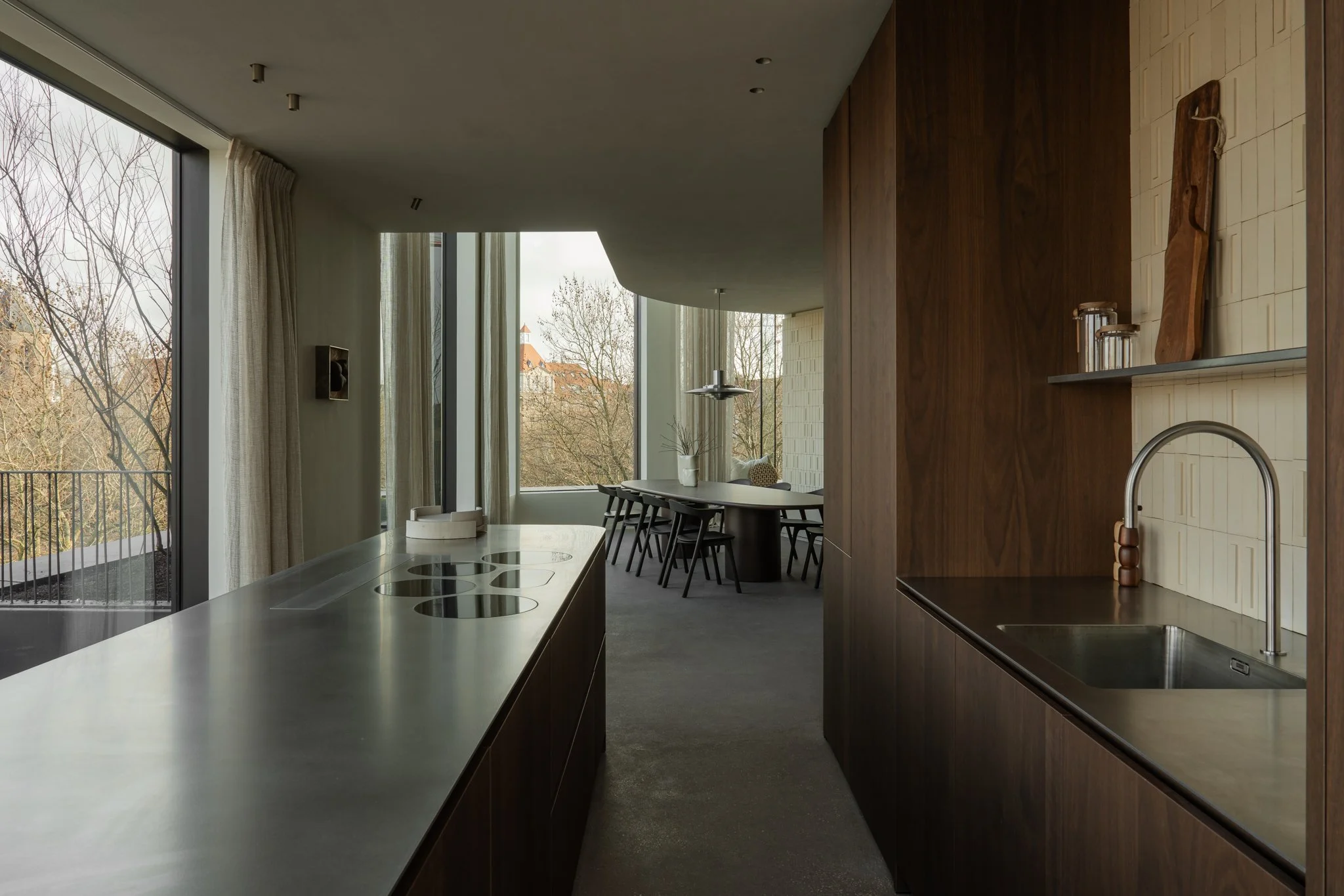 Modern kitchen with dark wood cabinets, a stainless steel sink, and a large window overlooking trees and buildings, with a dining area in the background.