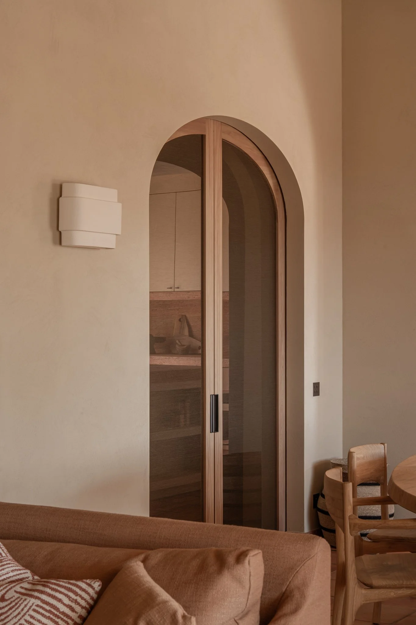 Interior view of a room featuring a wooden arched sliding door with glass, a beige sofa with cushions, a wooden dining chair, a bucket basket, and a modern wall-mounted light fixture.
