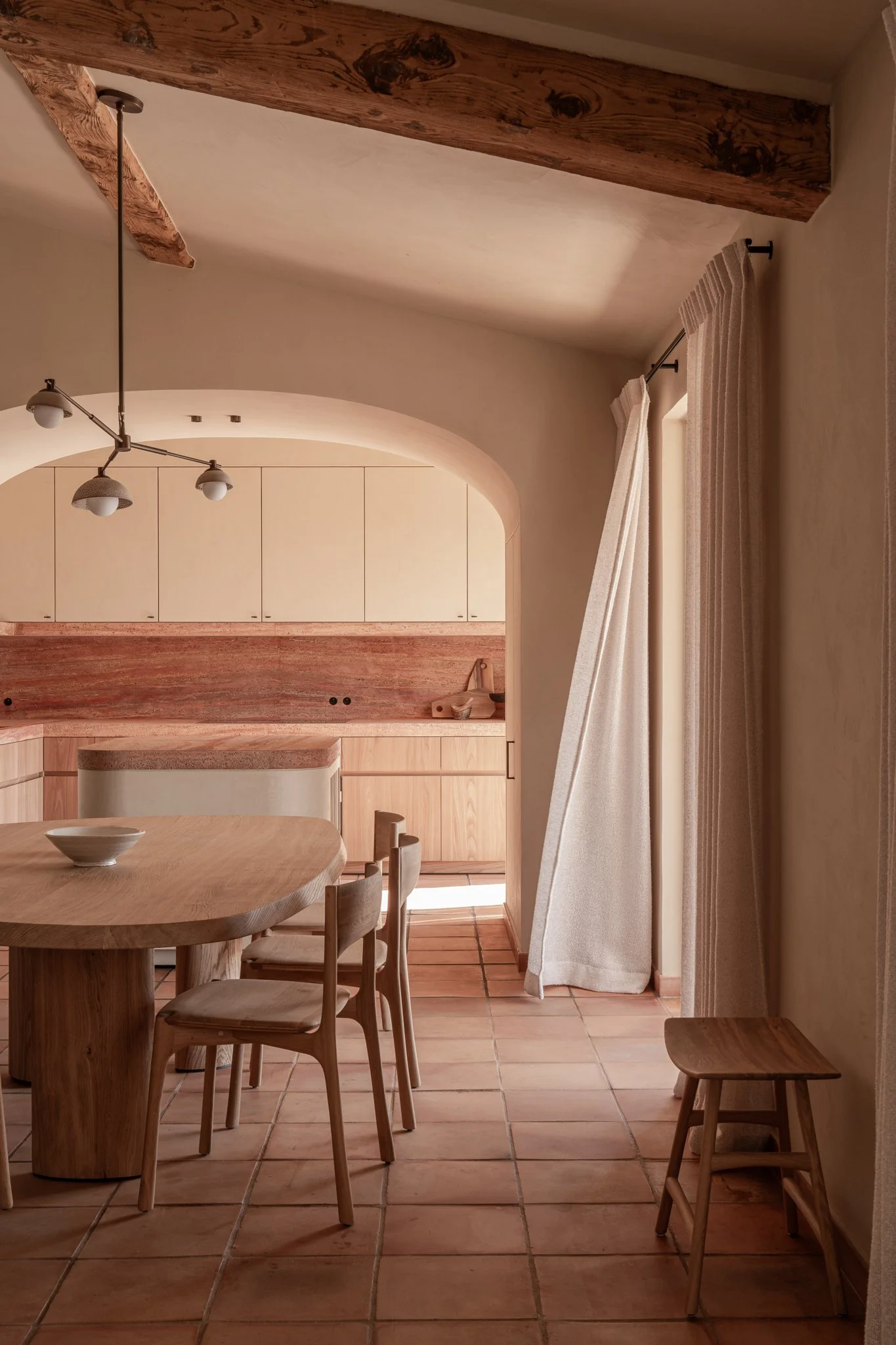 A cozy kitchen and dining area with wooden furniture, beige cabinets, terracotta tile flooring, and a curtain-covered door leading outside.