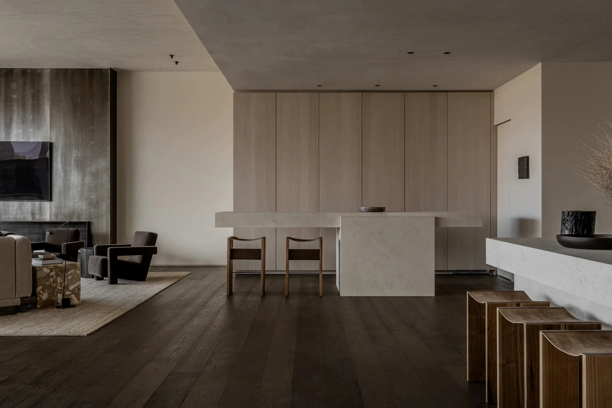 Modern living room with dark hardwood floors, a beige sofa, black armchairs, a mounted flat-screen TV, a textured wall, and a minimalist kitchen with light wood cabinets and a white counter.