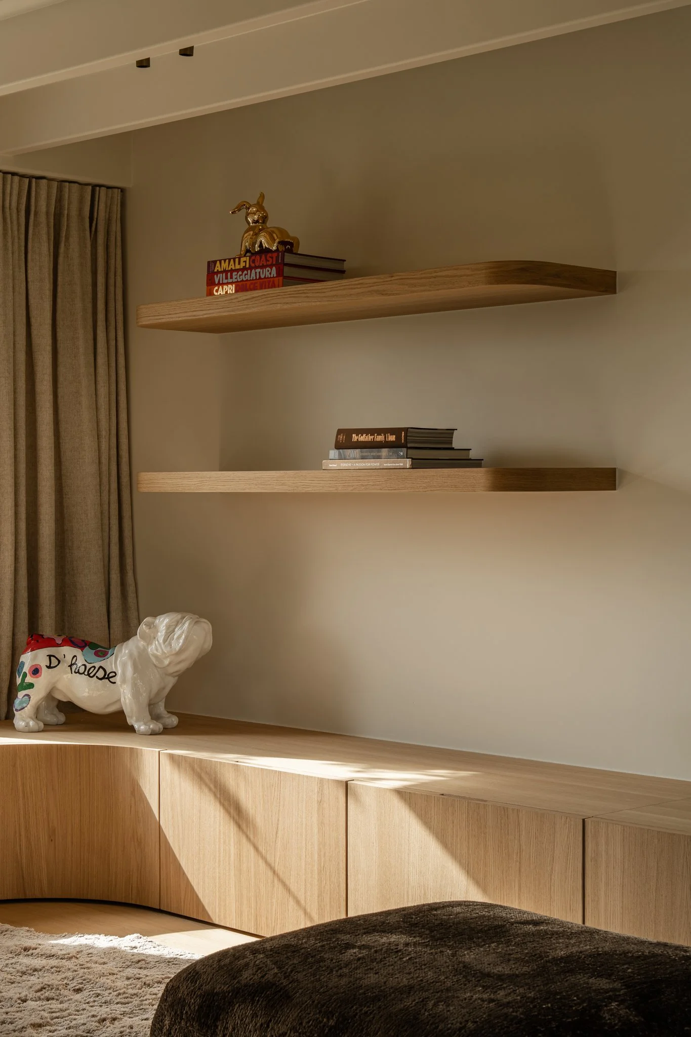 A living room with two wooden floating shelves on a beige wall, a ceramic dog sculpture on a wooden cabinet, and a partially visible dark brown upholstered piece of furniture.
