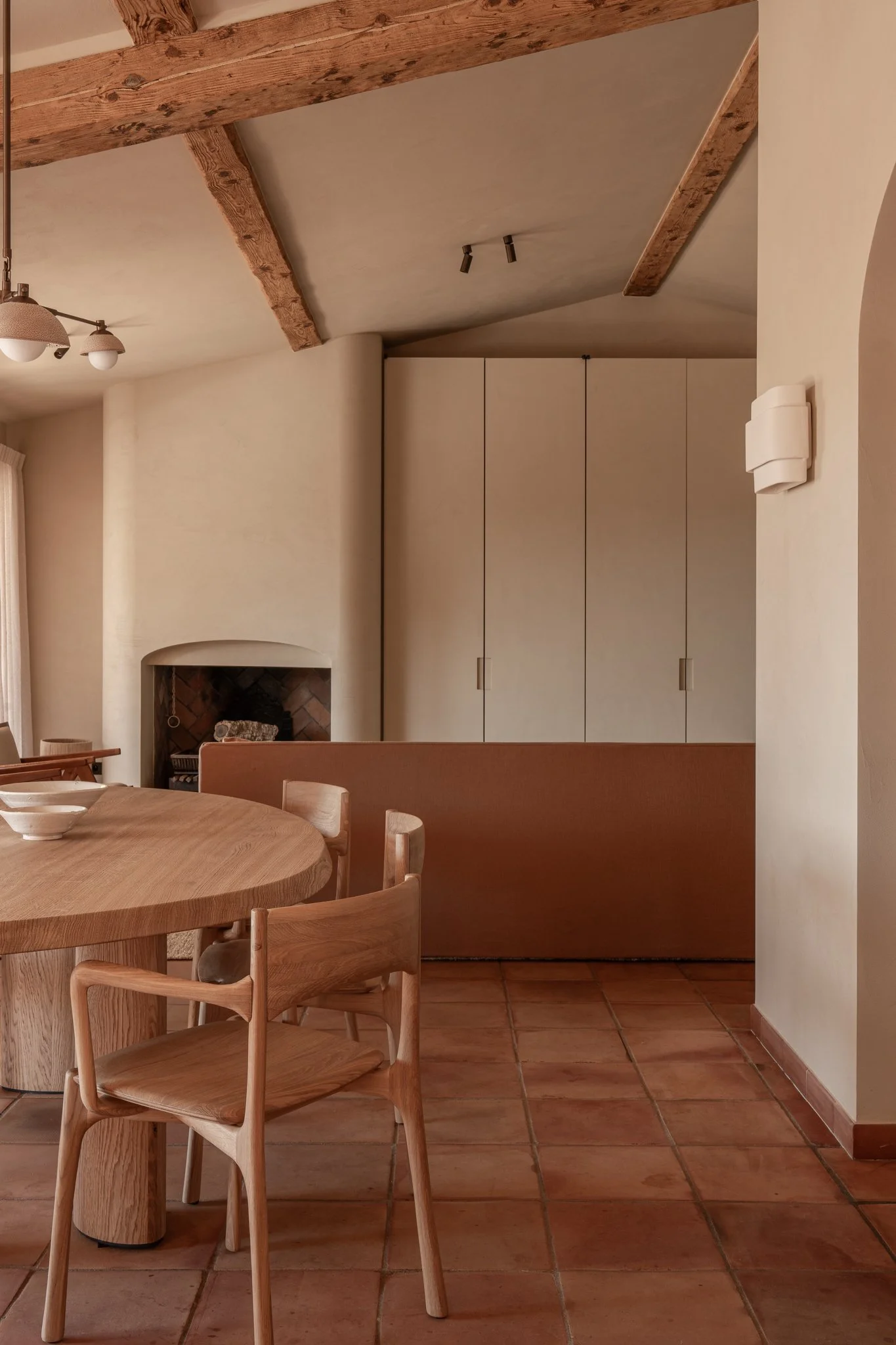 A cozy dining room with a wooden table and chairs, a fireplace, a built-in cabinet, and wood beams on the ceiling.
