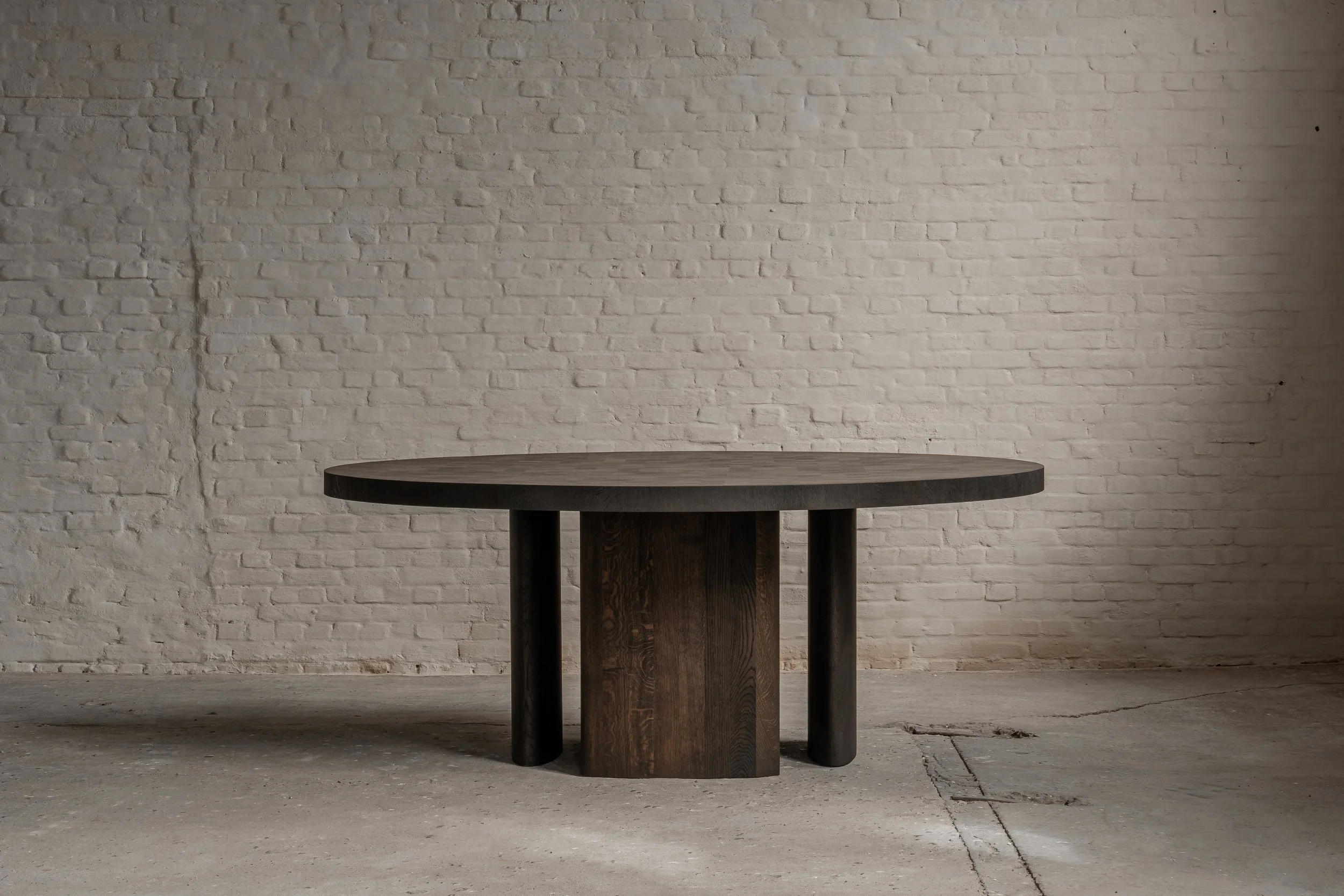 A round dark wooden table with three legs, positioned against a white brick wall and on a concrete floor.