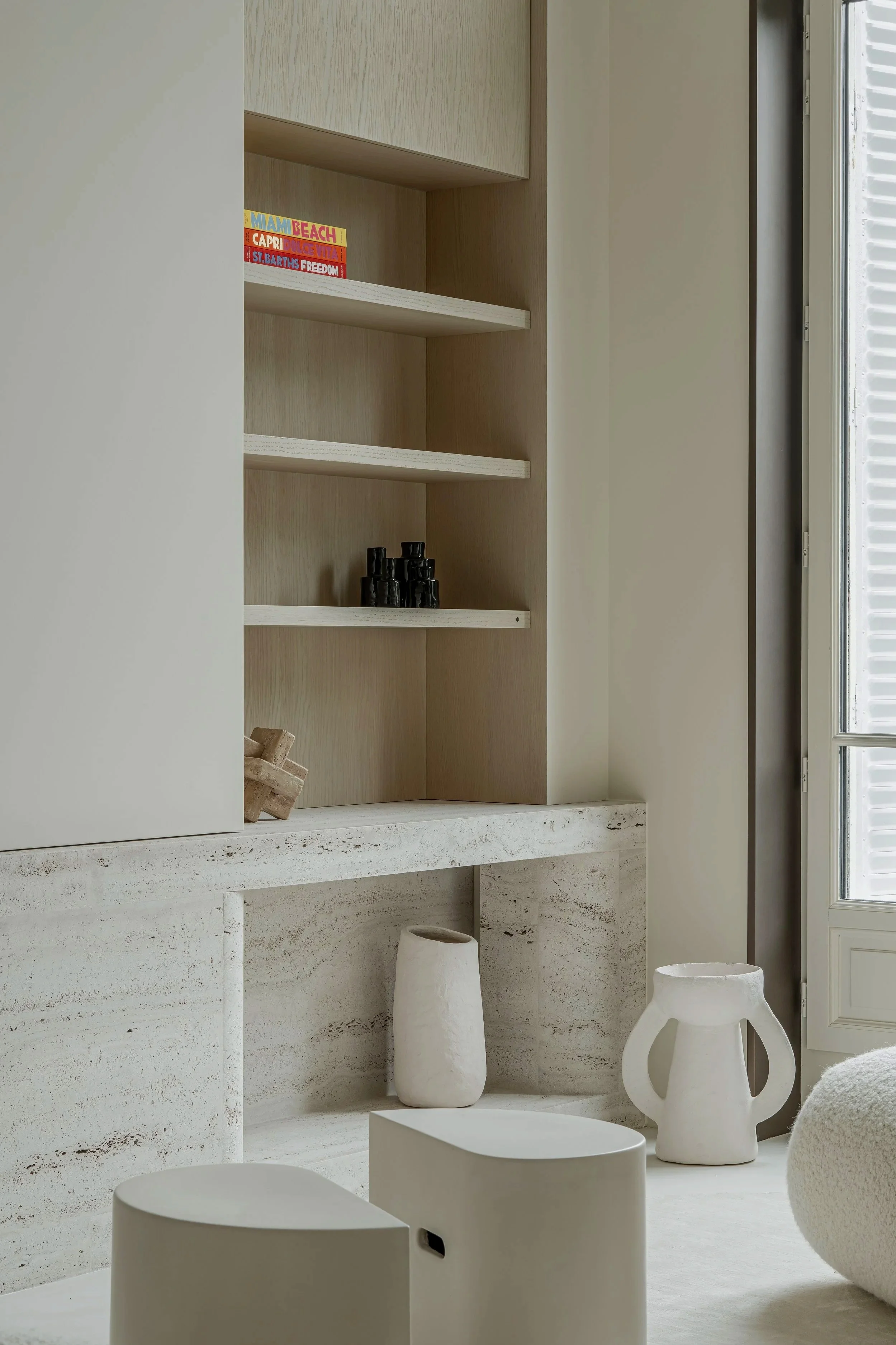 Minimalist interior with white and beige tones, featuring a built-in wooden and stone shelf with decorative pottery and small objects, next to a window with shutters.