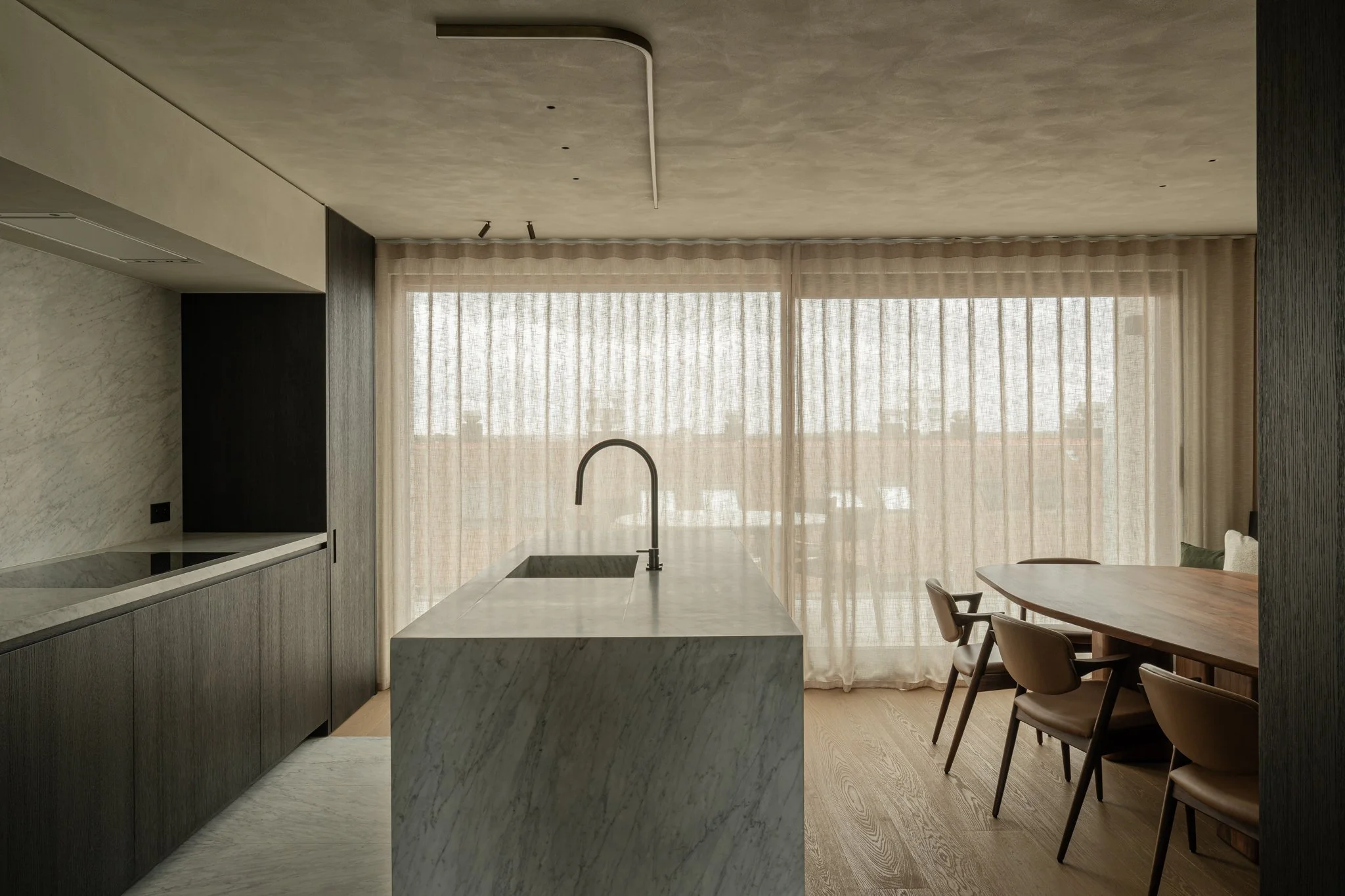 Modern kitchen with marble island, beige curtains, wooden dining table, and chairs in a minimalist style.