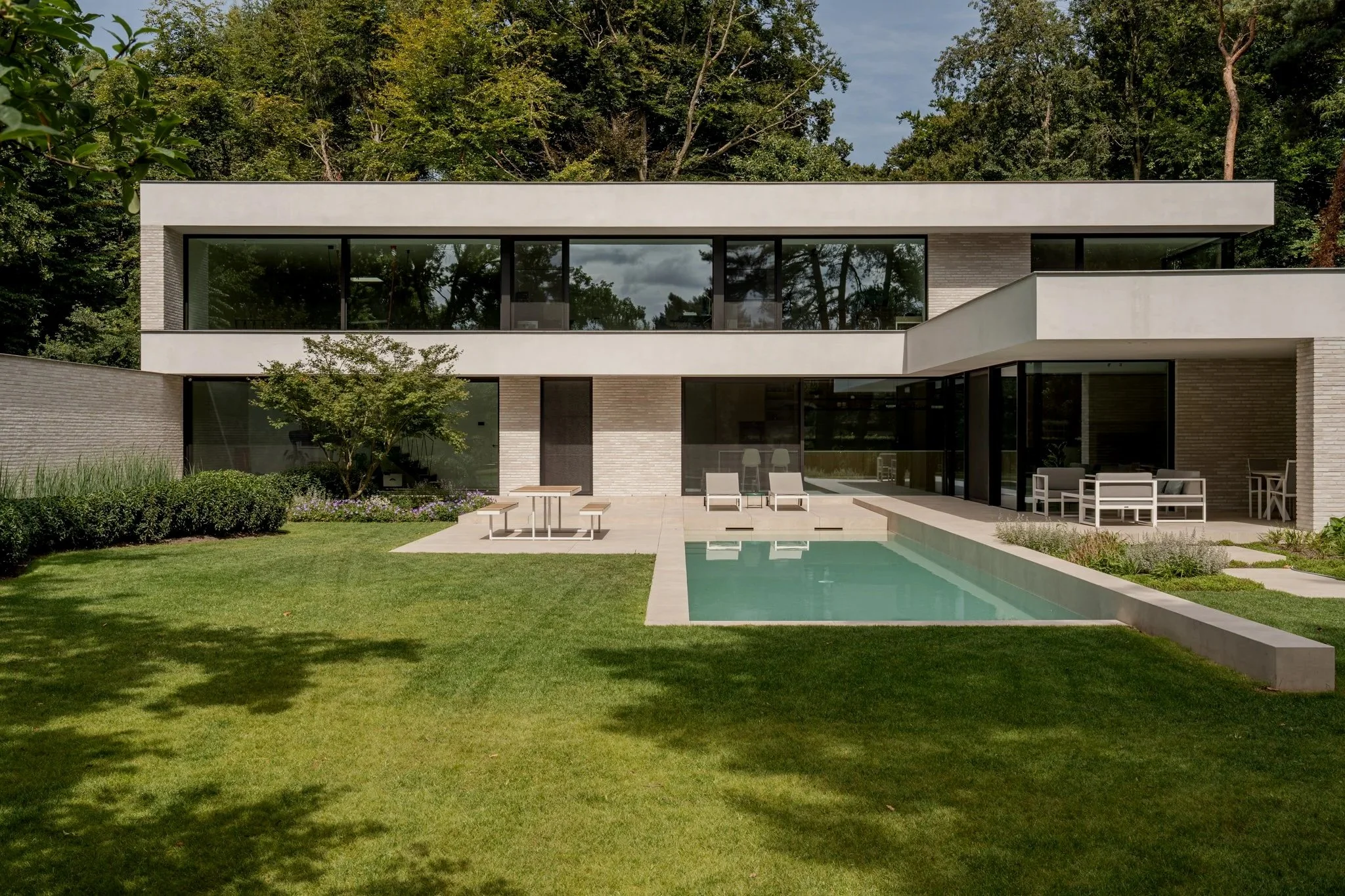 Modern two-story house with large glass windows, a lawn, pool, lounge chairs, and outdoor dining area, surrounded by trees.