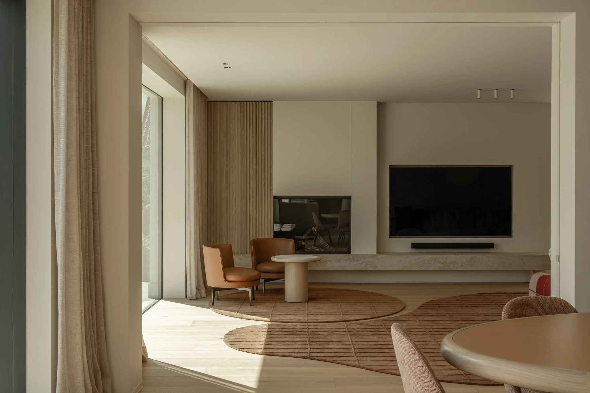 Modern living room with a flat-screen TV, two beige chairs, a small round table, and a fireplace with a glass cover. Light streams in through large glass doors, and the room features neutral tones and wooden accents.