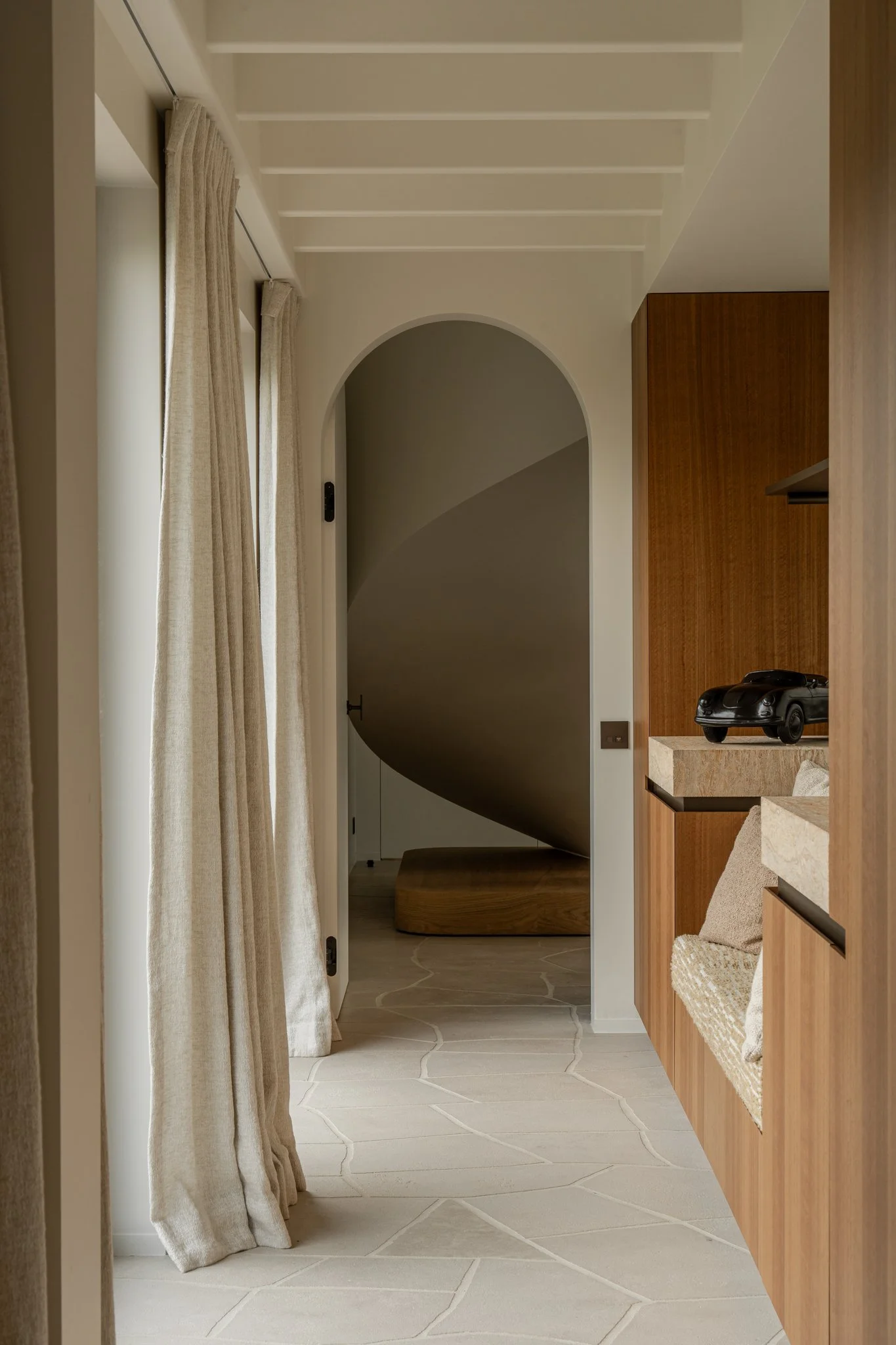 An interior hallway with cream-colored curtains and a stone tile floor leading to a rounded archway with a spiral staircase.