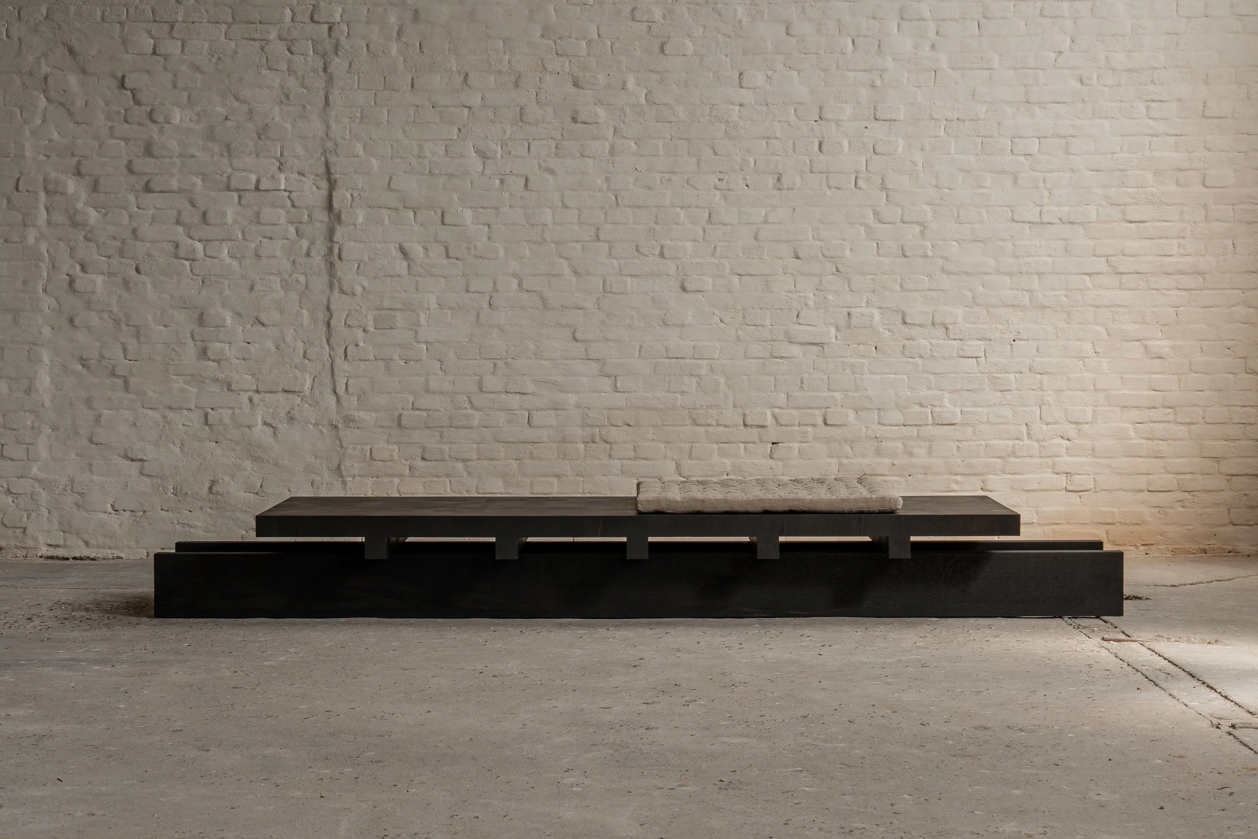 A low black wooden platform or bench against a white brick wall, with a rolled-up fabric or mat placed on top.