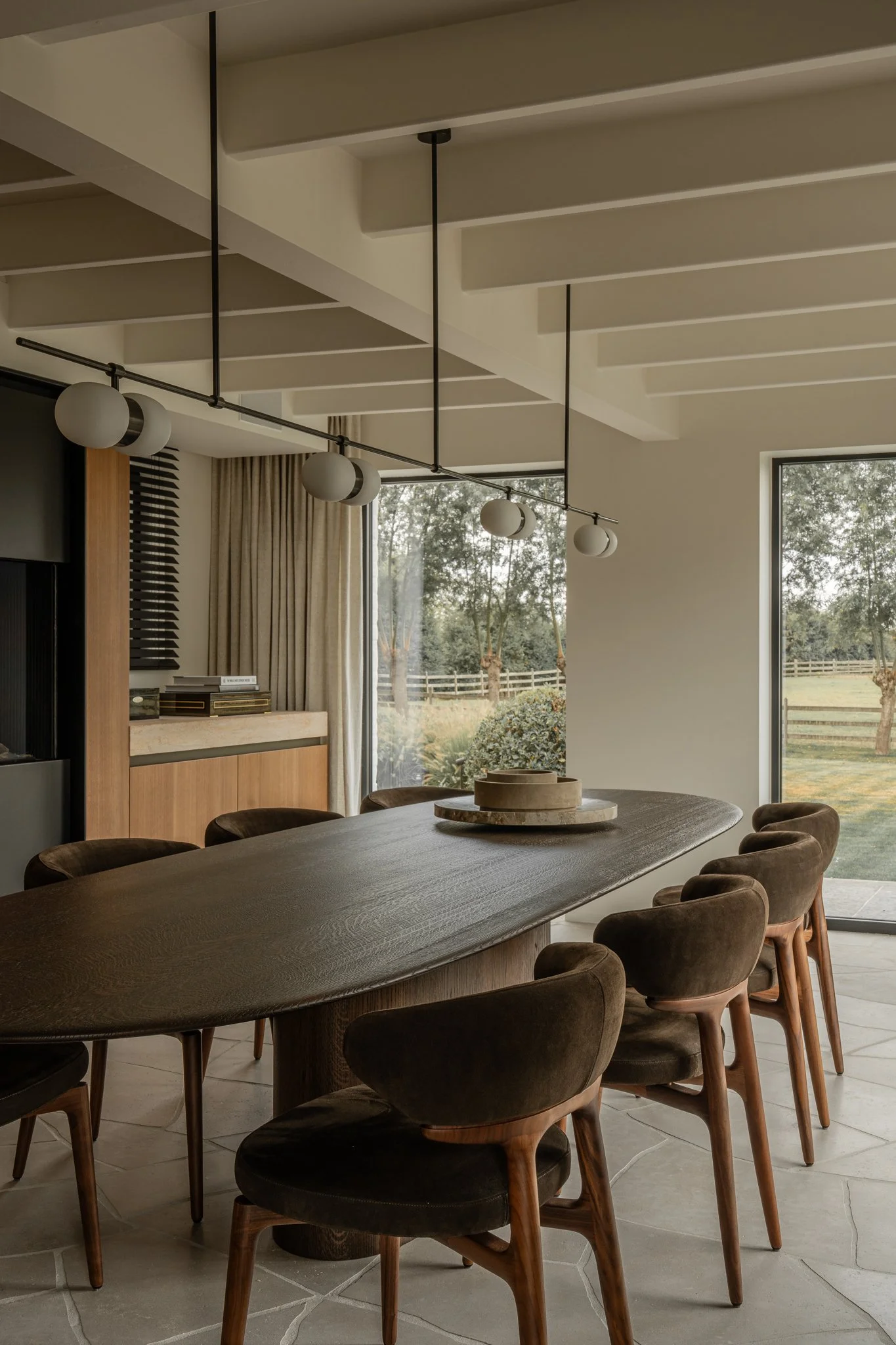 Modern dining room with a large wooden table, eight upholstered chairs, a contemporary chandelier, and large windows showing an outdoor garden area.