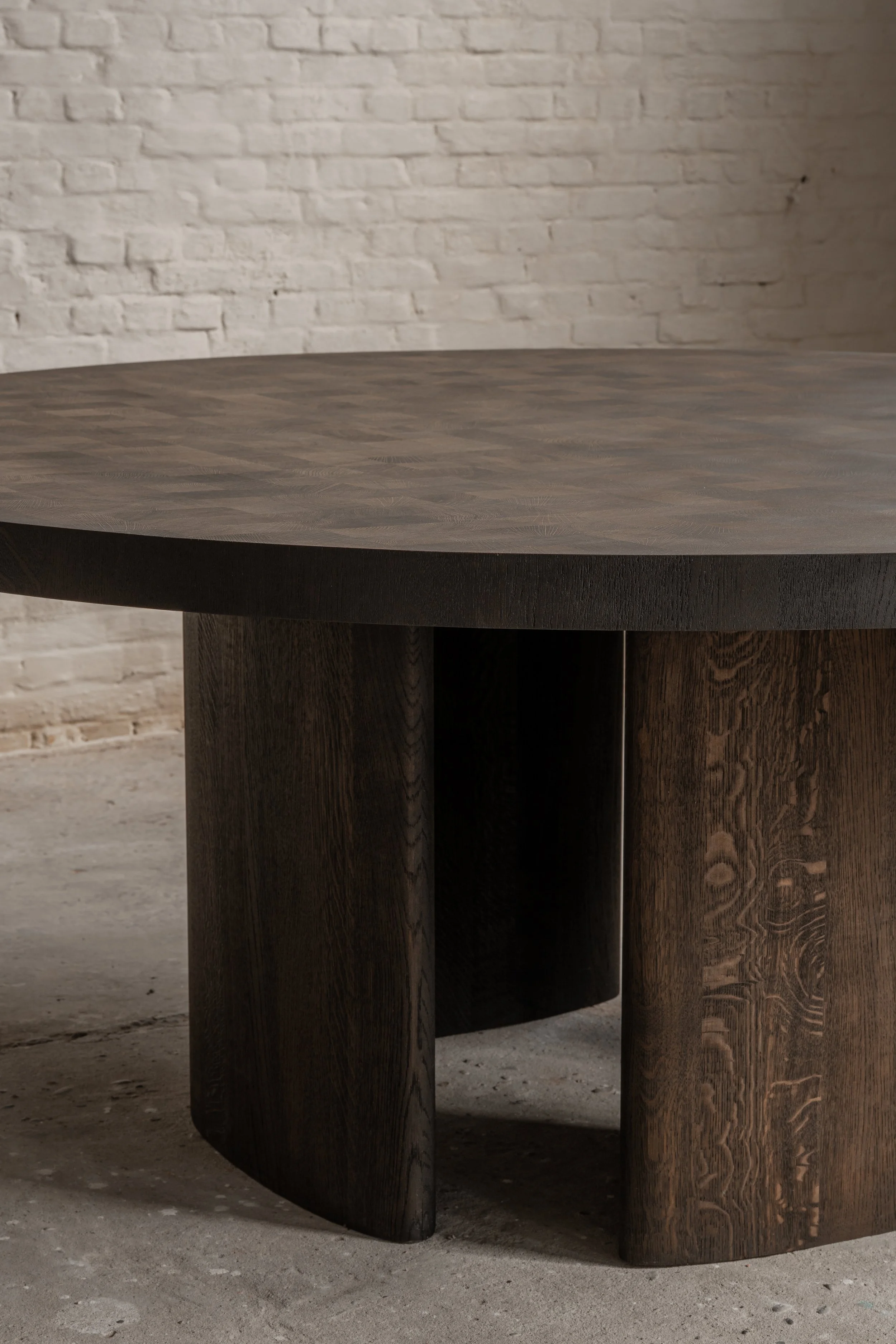 Close-up view of a round, dark wooden table with a thick, sturdy base, positioned on a concrete floor with a white brick wall in the background.