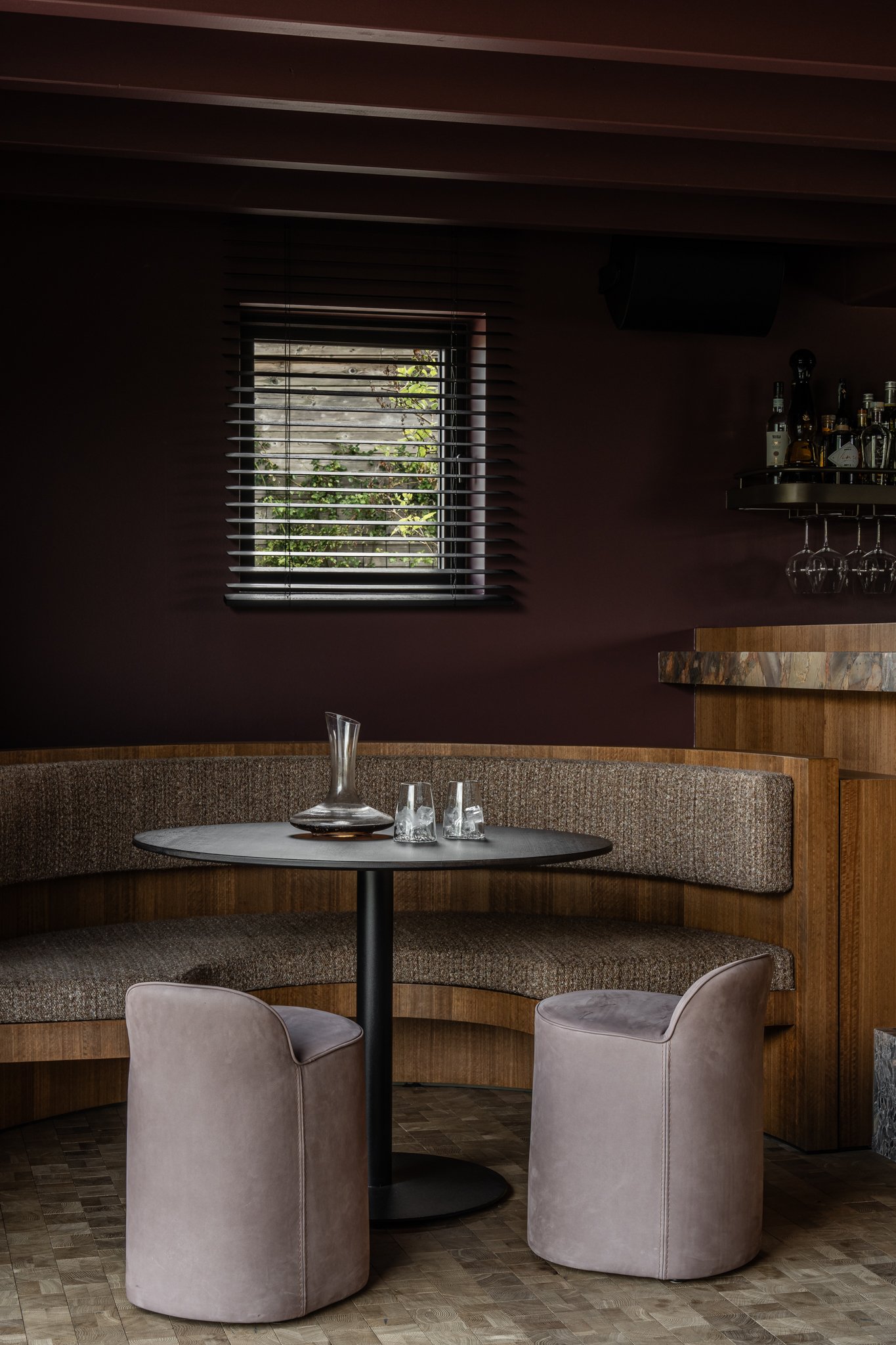 Empty seating area with a round table, a curved bench, and two cushioned chairs, set near a window with blinds, in a dimly lit room.