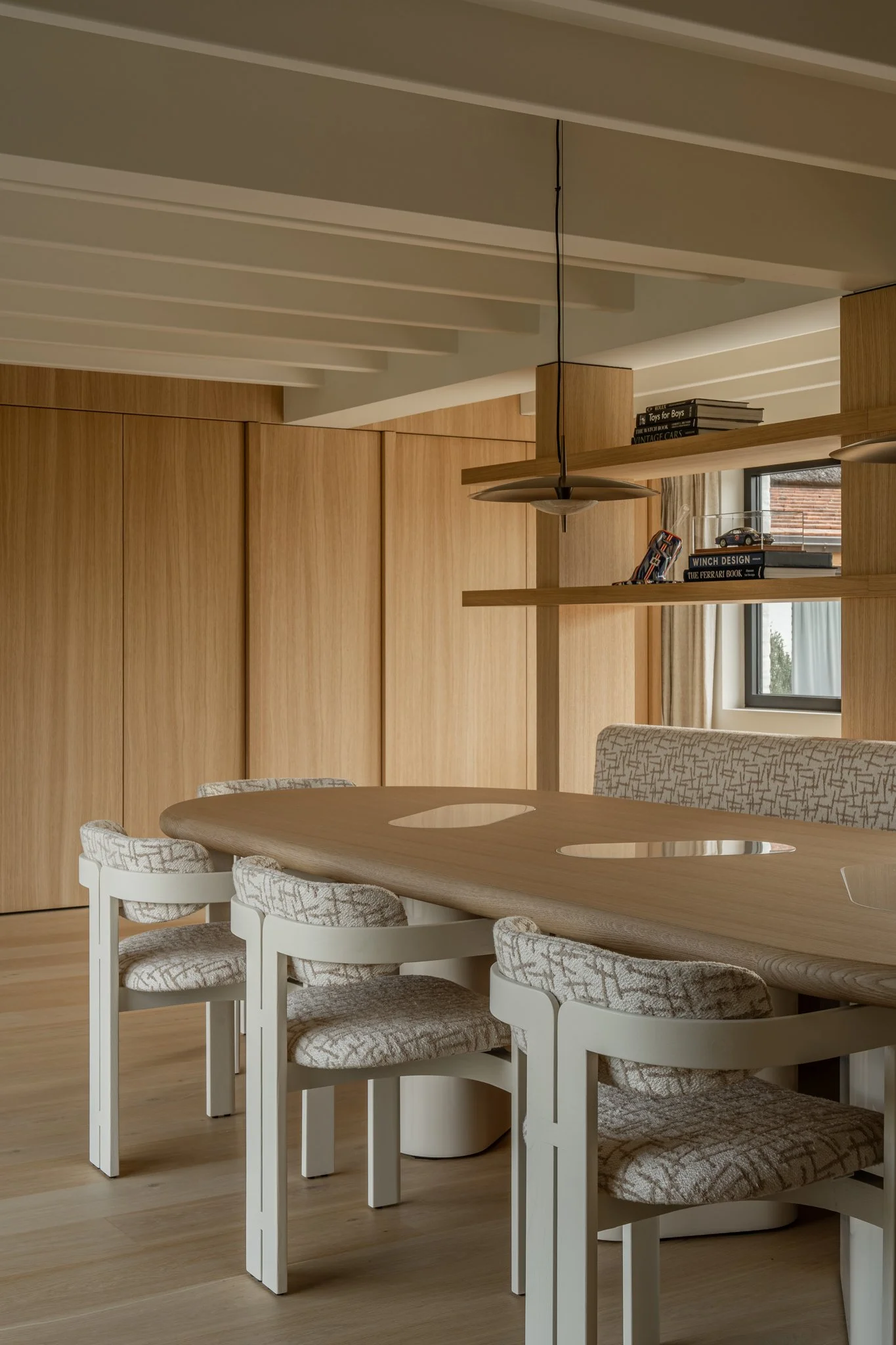 Modern dining room with a wooden table, upholstered chairs, wood paneling on the walls, and shelves with books and model cars, illuminated by natural light from a window.