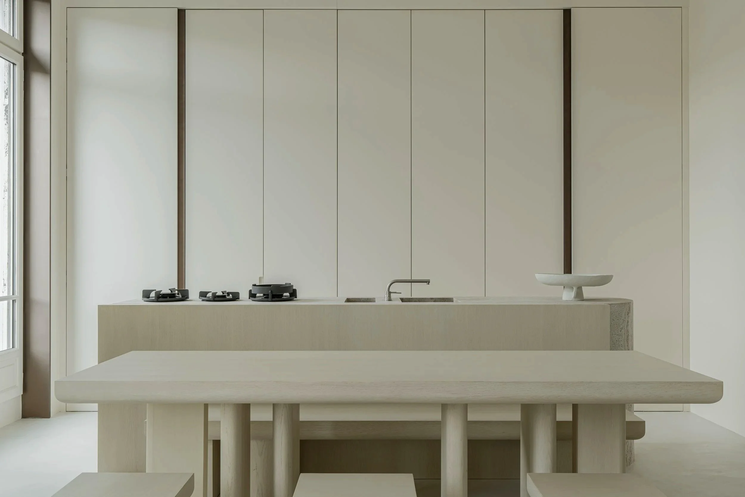 Minimalist kitchen with a light wood dining table and benches, beige cabinets, black stove burners, a sink with a faucet, and a decorative white bowl on the counter. Large window on the left wall.