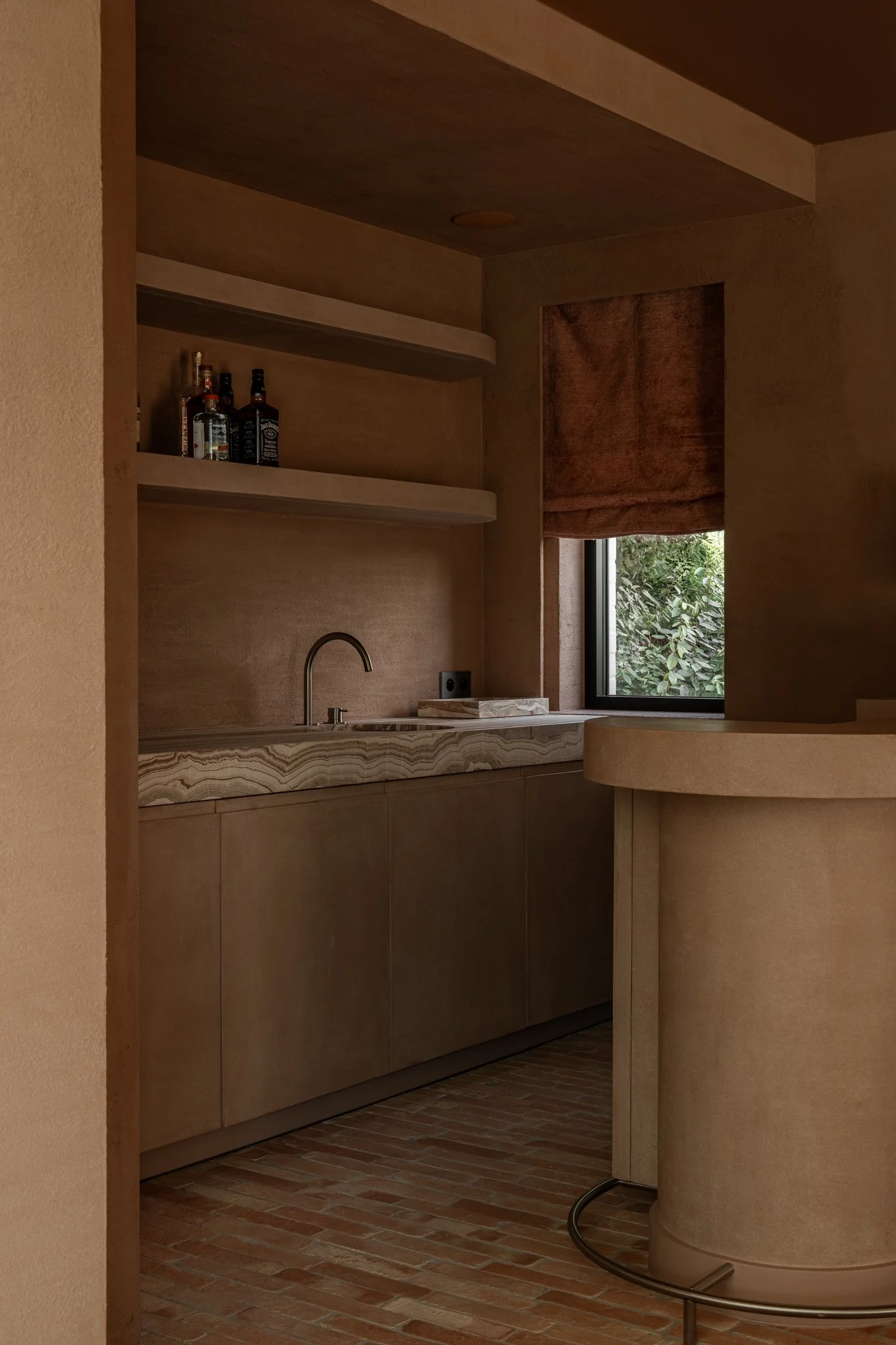 A small kitchen with beige walls, a window with a brown curtain, a marble countertop, and a few bottles on a shelf above. There is a faucet on the counter and a piece of paper or napkin next to it.