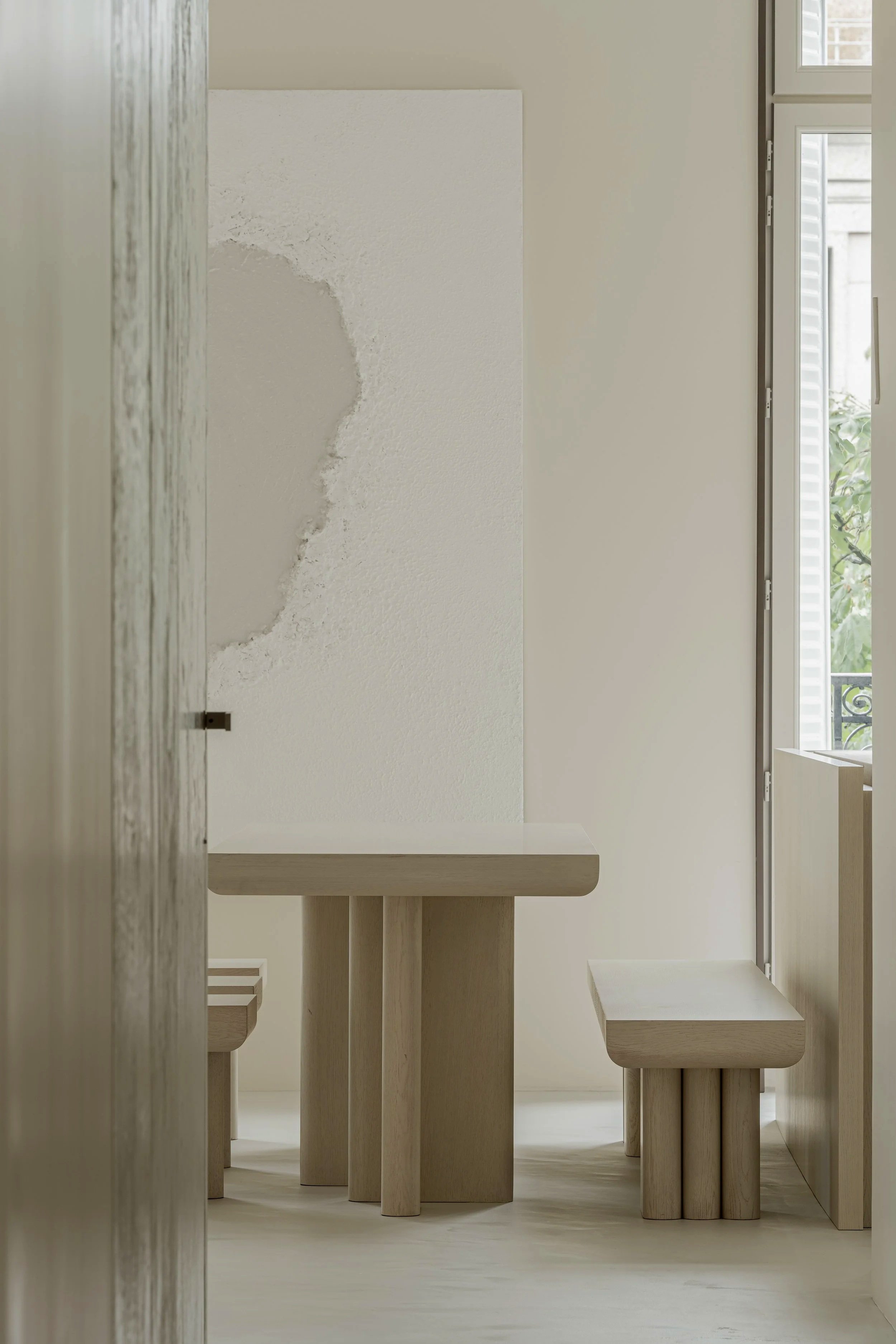 Minimalist interior with wooden table and benches near a window, with a rough patch on the white wall.