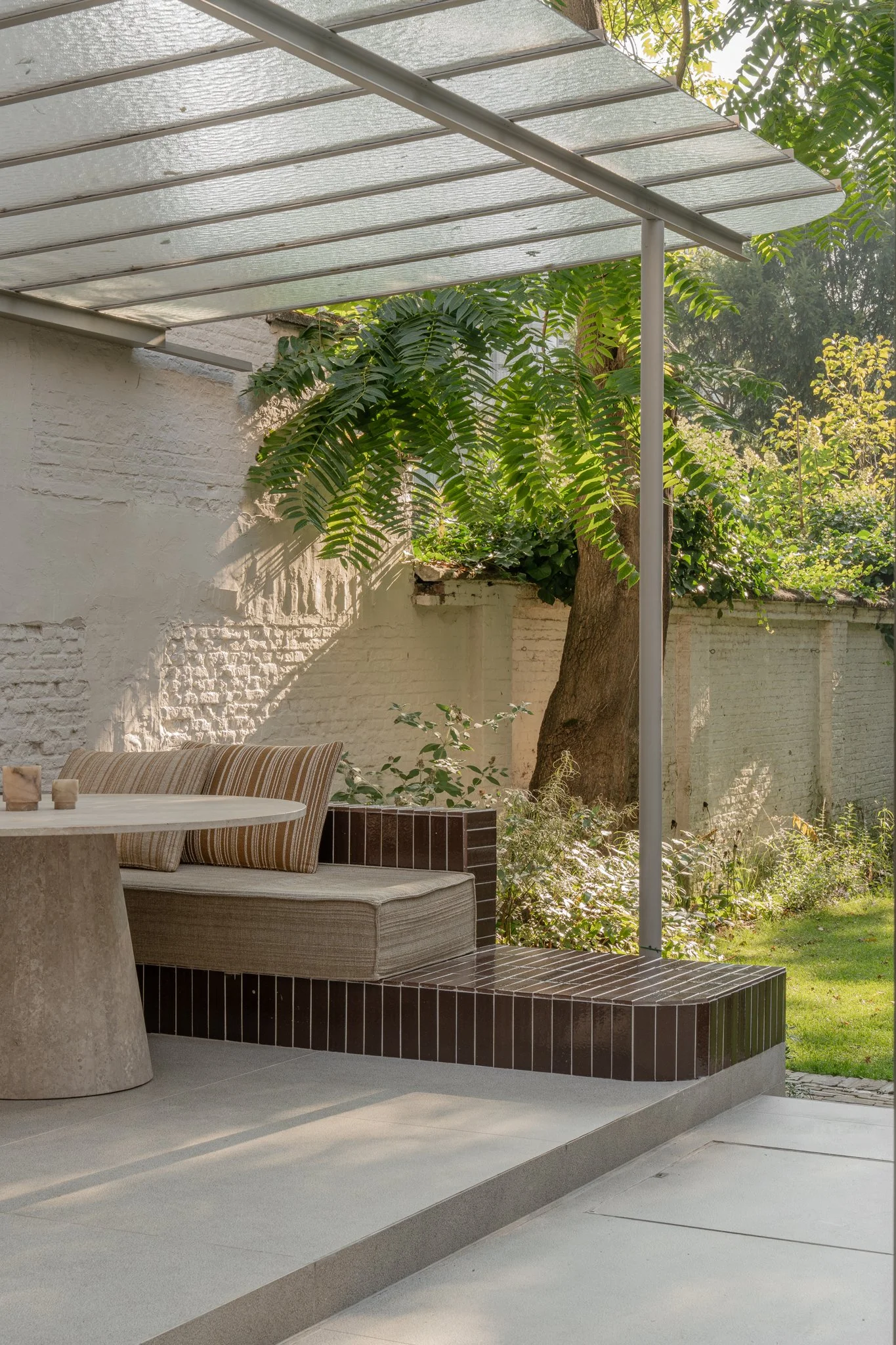 Outdoor patio area with a cushioned bench, round table, and partial view of a garden with trees and greenery.
