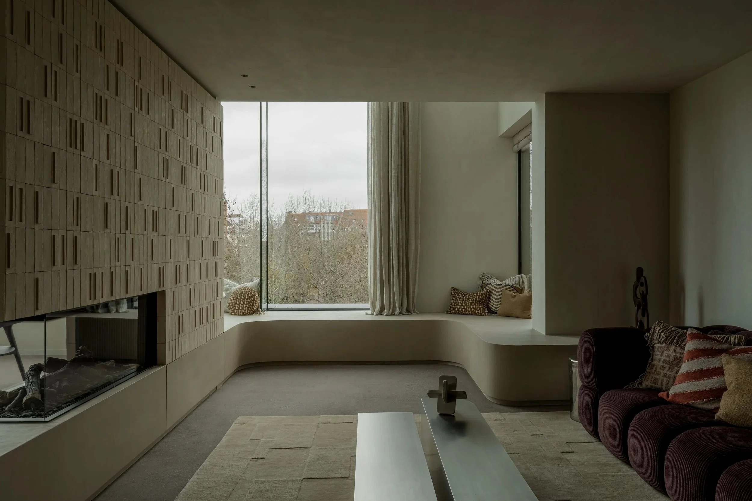 Modern living room with a large window, window seat with pillows, and a built-in fireplace with a brick accent wall.