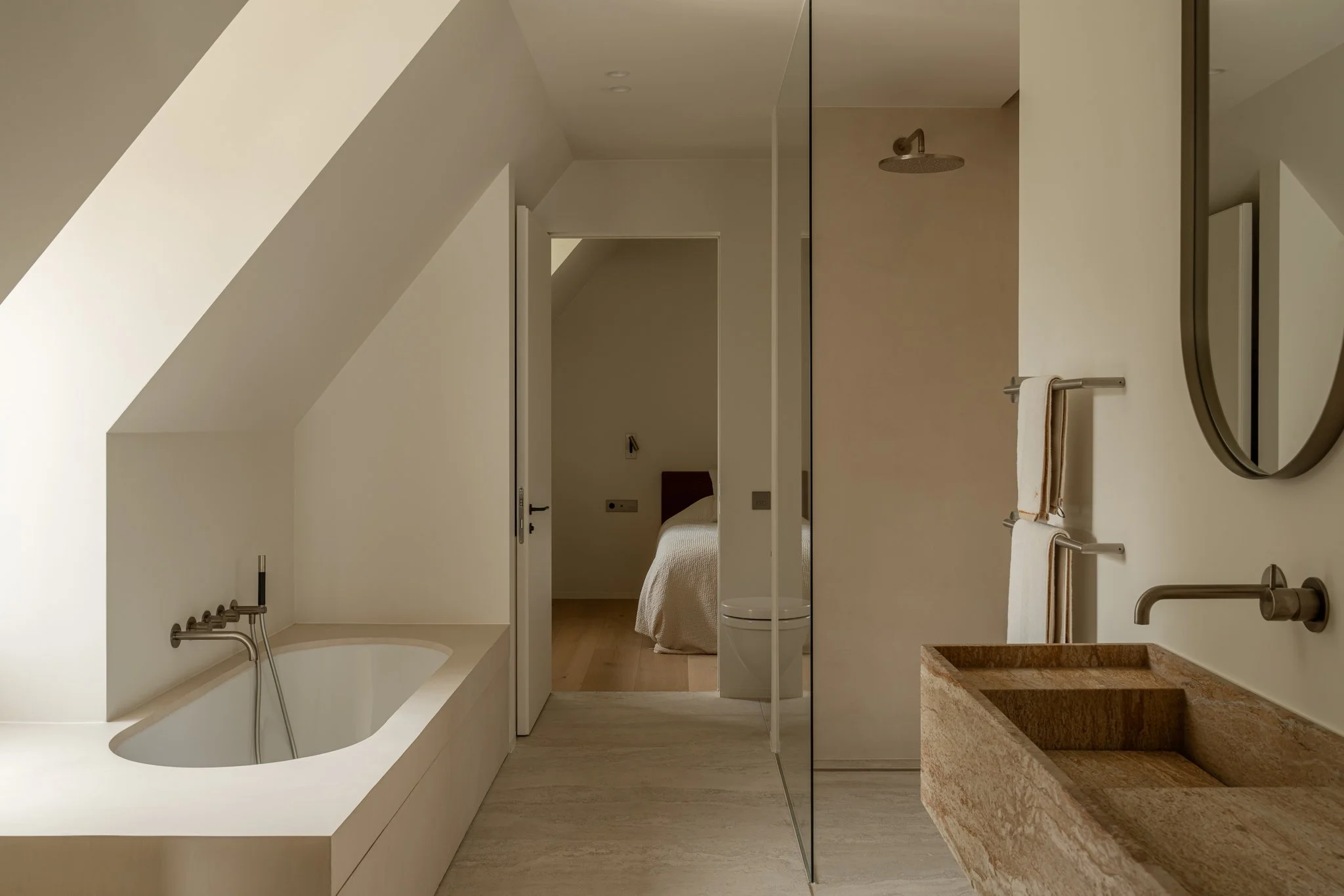 Modern bathroom with a built-in bathtub under the sloped ceiling, a stone sink with a wall-mounted faucet, a towel rack, and a shower area with a ceiling-mounted showerhead. Visible doorway leading to a bedroom with a bed.