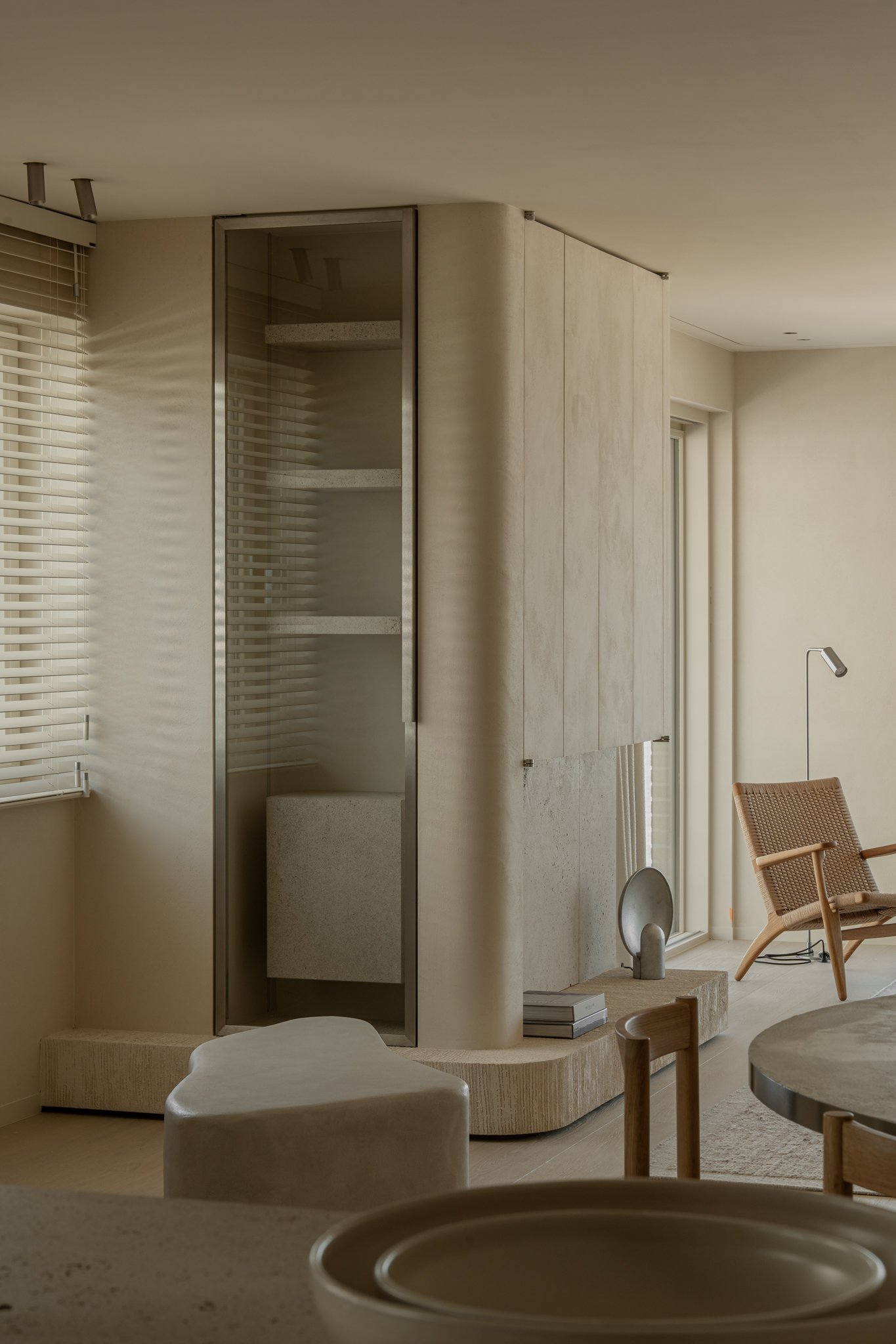 Interior of a modern living space with beige tones, a glass shower enclosure, a wooden chair, a reading lamp, and decorative objects.