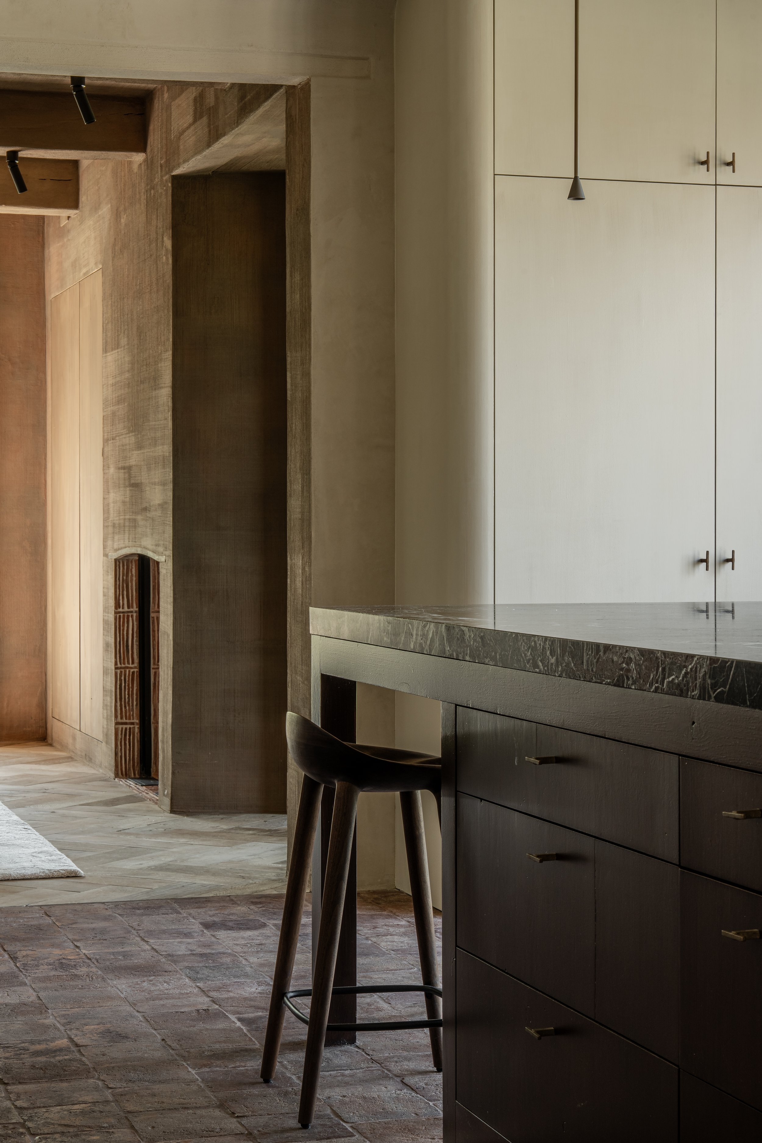 A modern kitchen with a black barstool, dark cabinetry, a marble countertop, and neutral-colored walls with a wooden door and cabinets in the background.