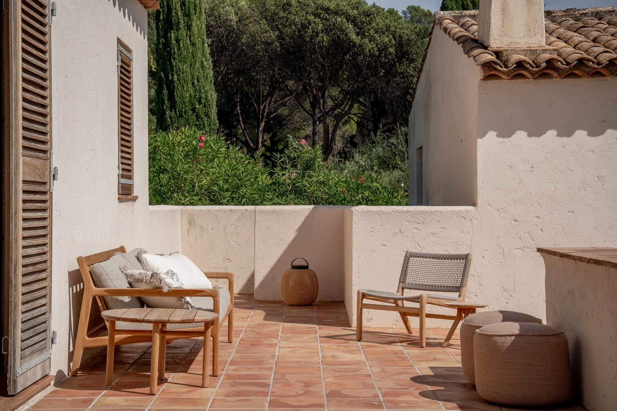 An outdoor terrace with terracotta tile flooring, beige stucco walls, and wooden furniture including a bench with pillows, a chair, and two rounded ottomans. There is a woven lantern, and in the background, lush greenery with trees and flowering bushes under a clear sky.