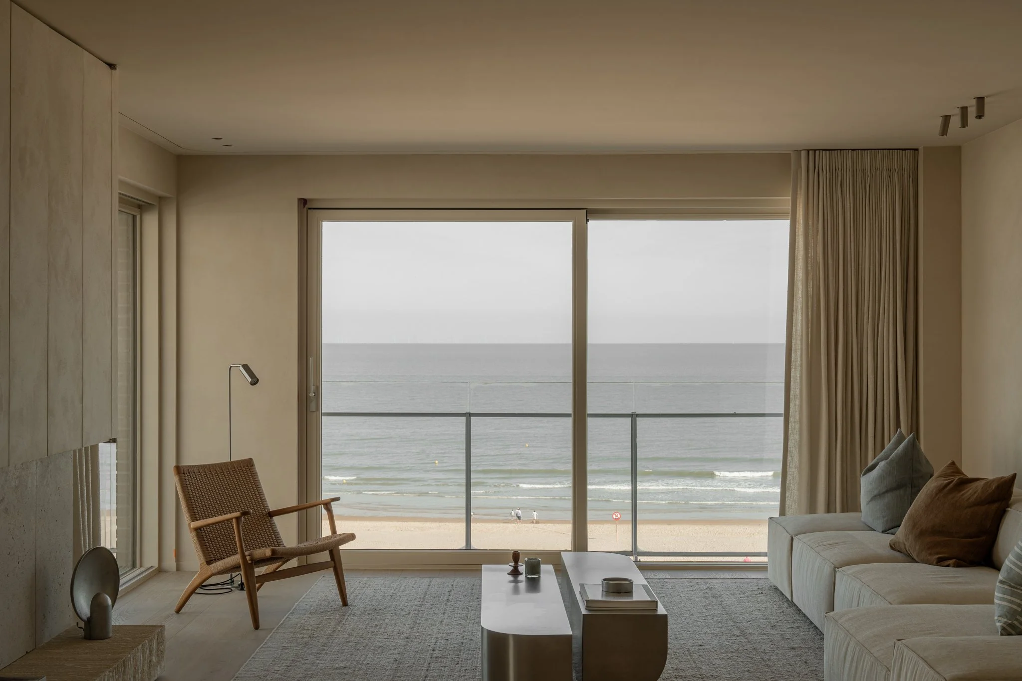 Living room with large sliding glass doors opening to a beach view, featuring a beige sofa with cushions, a white coffee table, a woven armchair, and minimal decor.