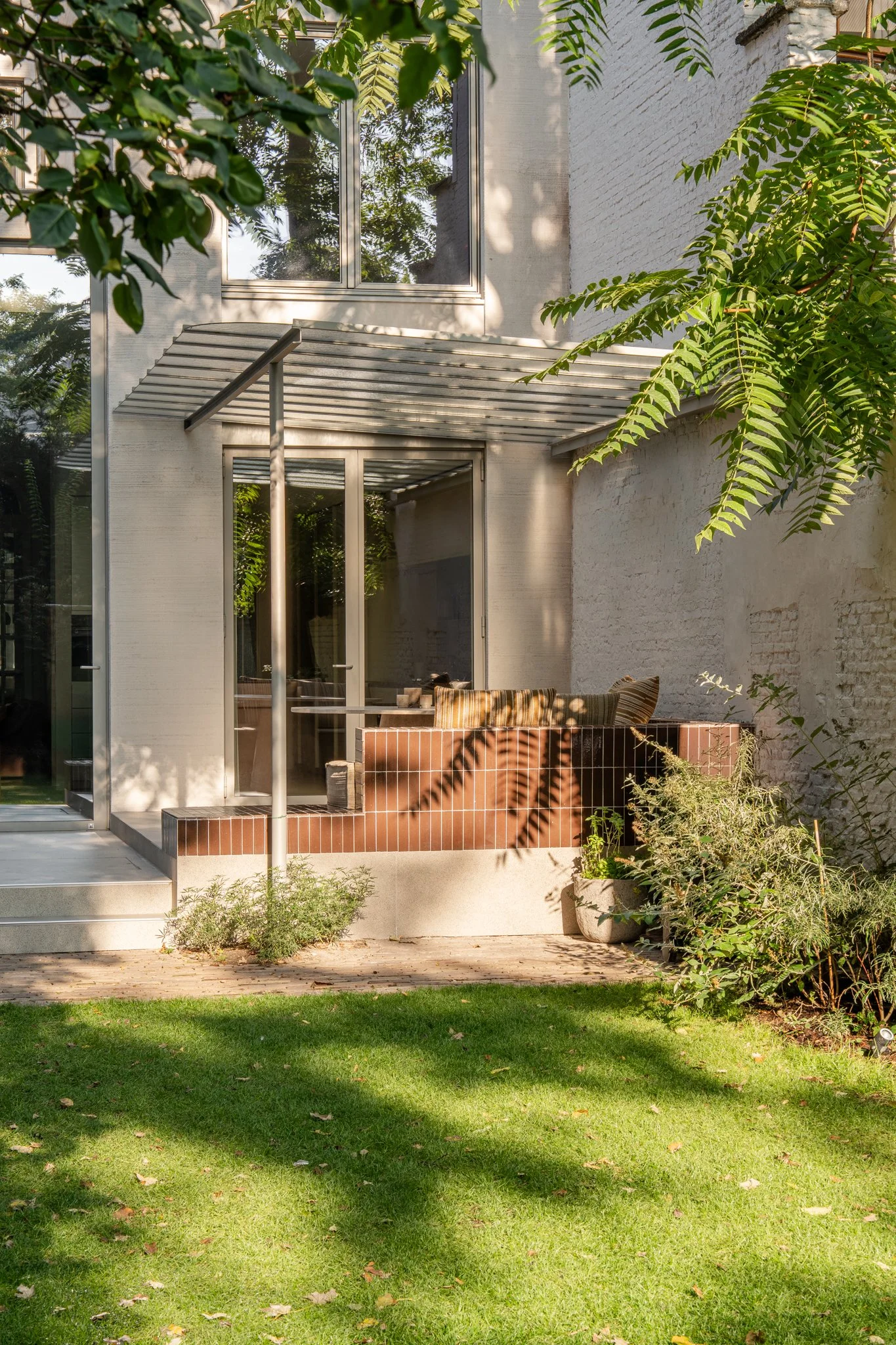 A modern house patio with sliding glass doors, a small overhang, and surrounding plants and greenery, with a grassy yard in the foreground.