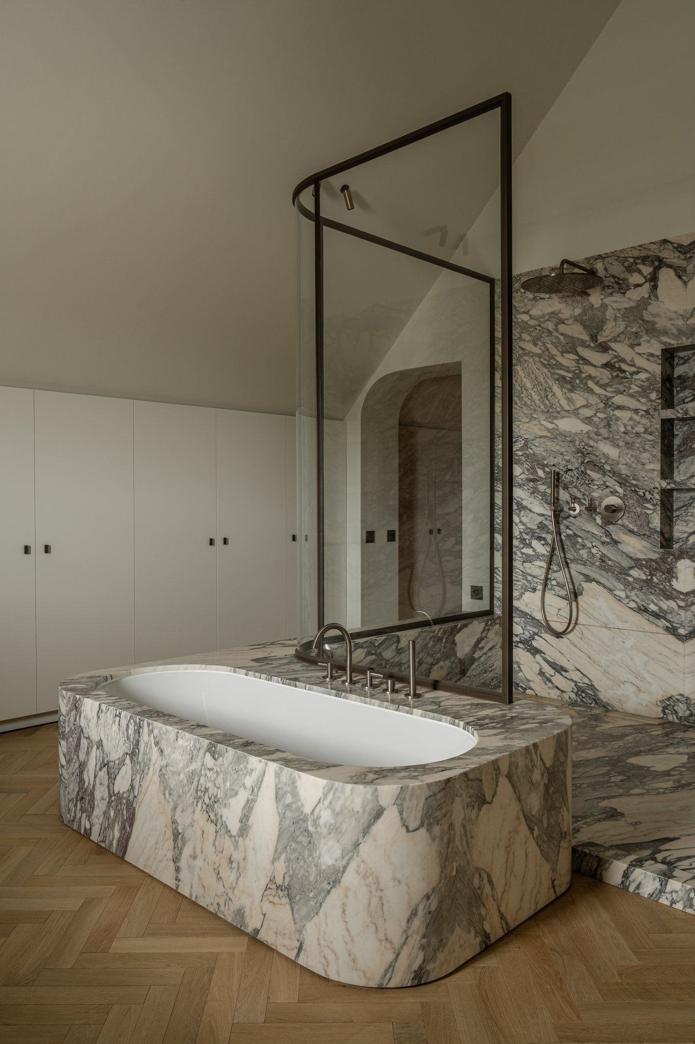 A modern bathroom featuring a marble bathtub, a large mirror, a marble shower wall with a rain shower head, and wooden flooring.