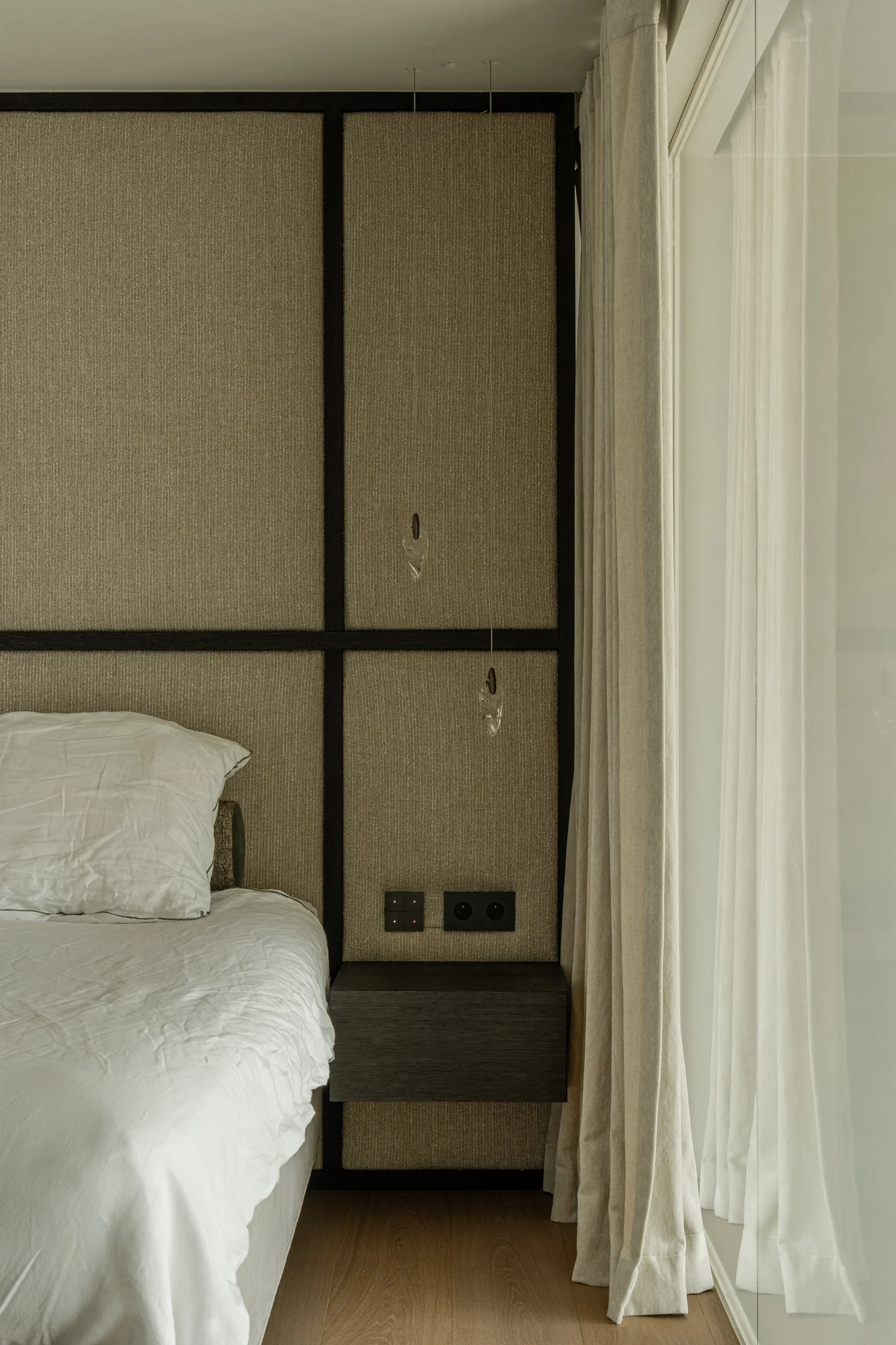 Close-up of a bedroom wall with electrical outlets, a small floating nightstand, and a beige curtain beside a glass window.