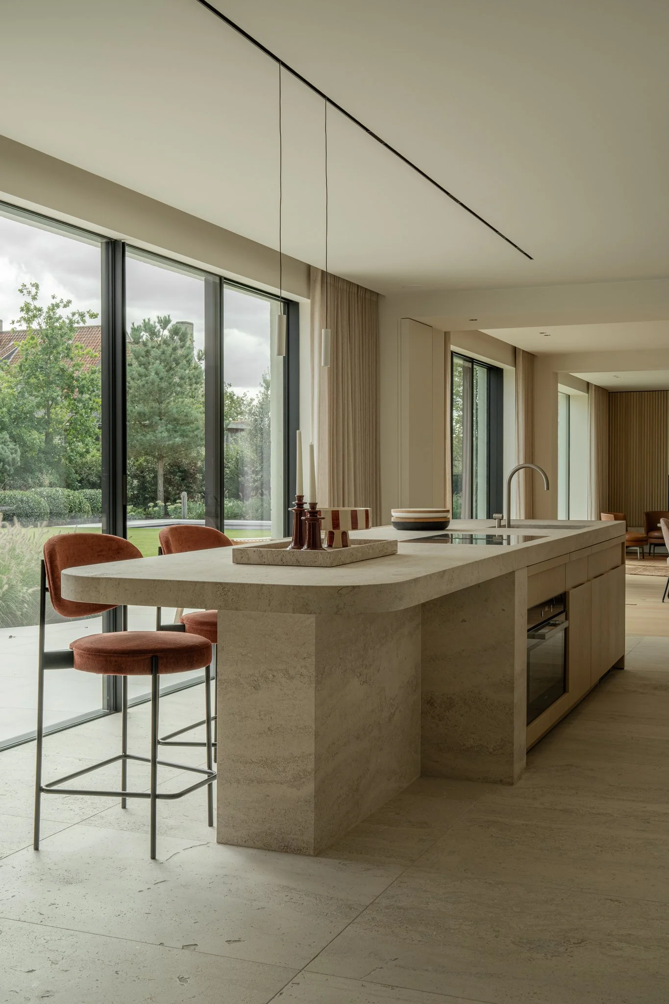 Modern kitchen with a curved stone island, two rust-colored bar stools, large windows showing outdoor greenery, and minimalistic decor.