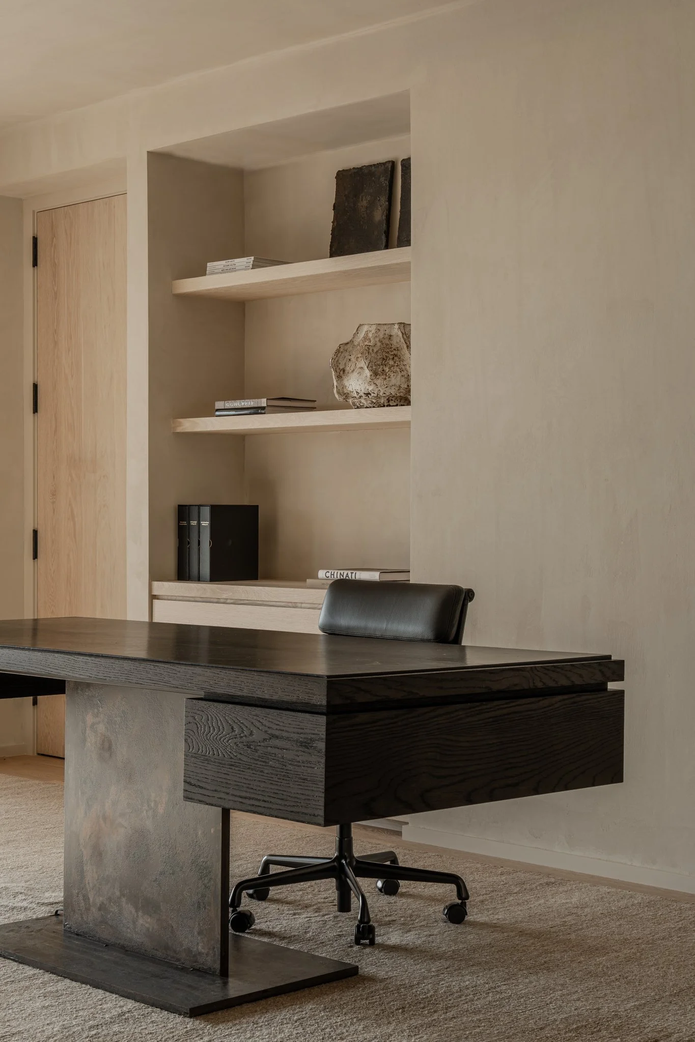 Modern black wooden desk with a swivel office chair in an office room, beige walls, and built-in shelves with books and sculptural objects.