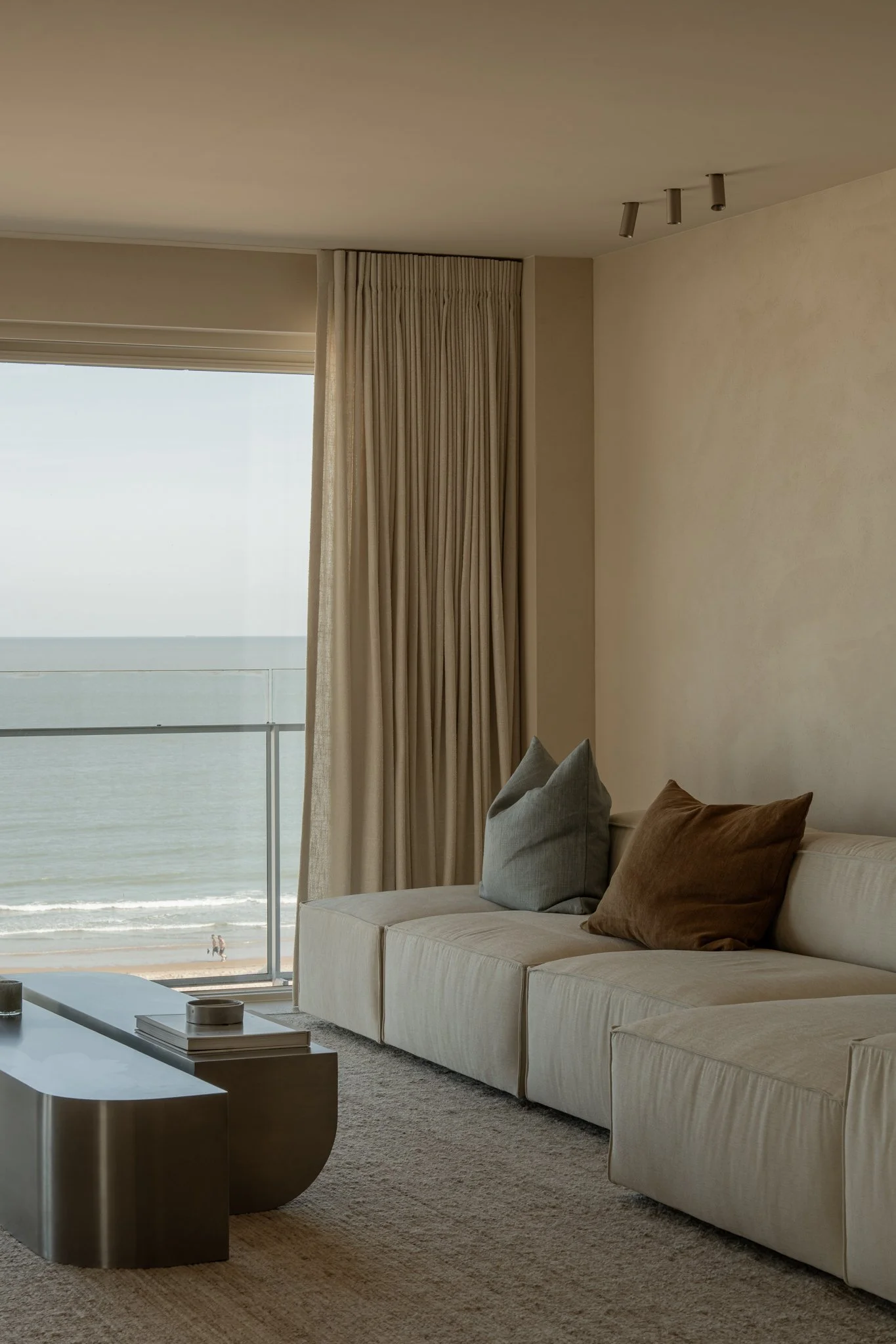 Living room with a large window overlooking the ocean, a cream sofa with two colored pillows, a modern metallic coffee table, and beige curtains.
