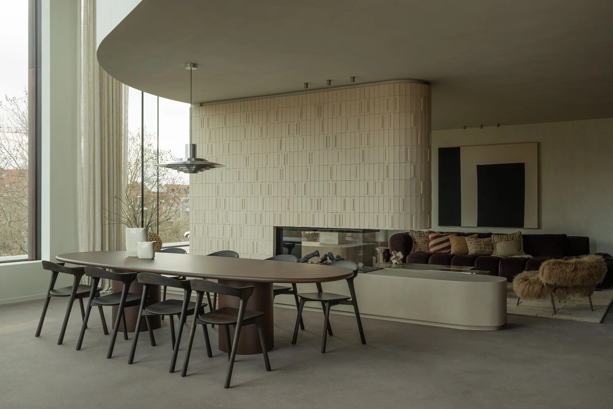 Modern living room with a large corner window, a dining table with six chairs, and a sofa with multiple pillows. There's a fireplace with a unique textured wall and a piece of modern art hanging above the sofa. A decorative plant and vases sit on the dining table, and a furry throw is on a chair near the sofa.