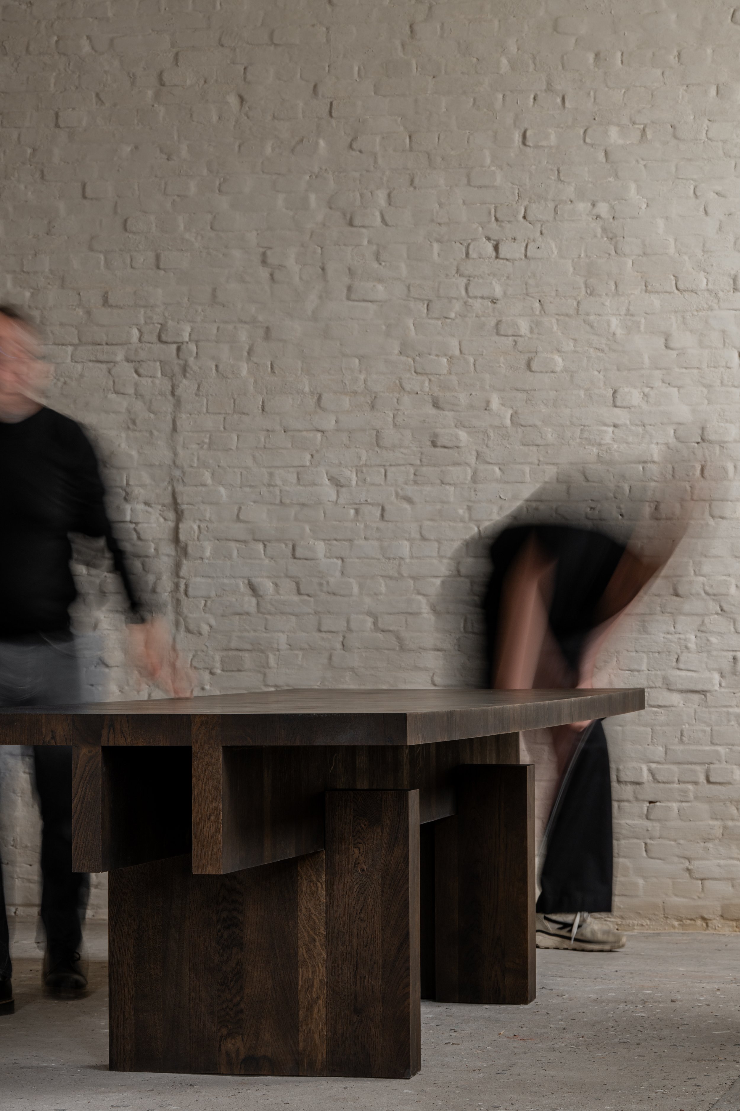 Two people are moving a large wooden table against a white brick wall, one person is blurred on the left and the other is behind the table on the right.