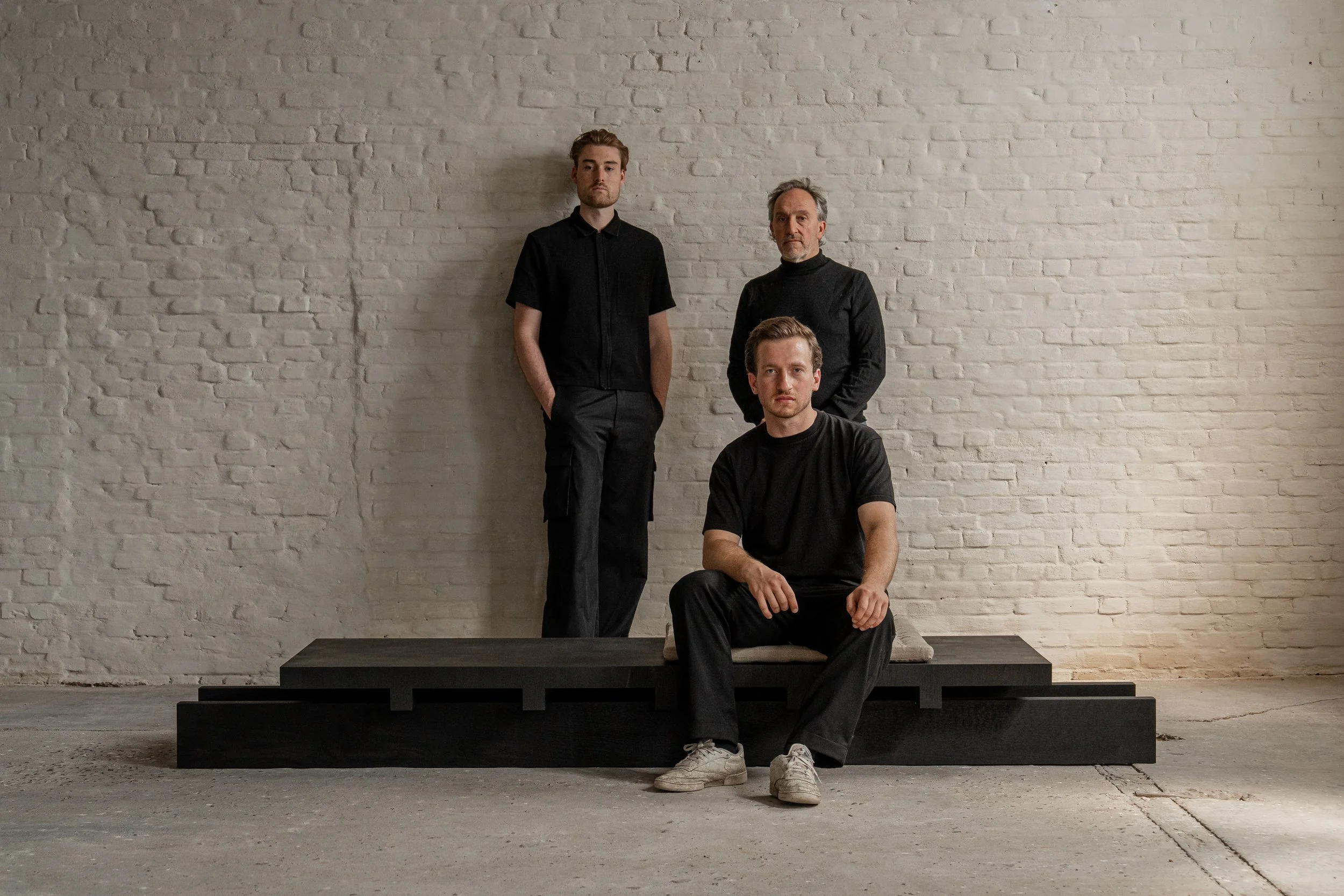 Three men, one sitting on a low platform and two standing behind him, pose against a white brick wall in a minimalistic setting.