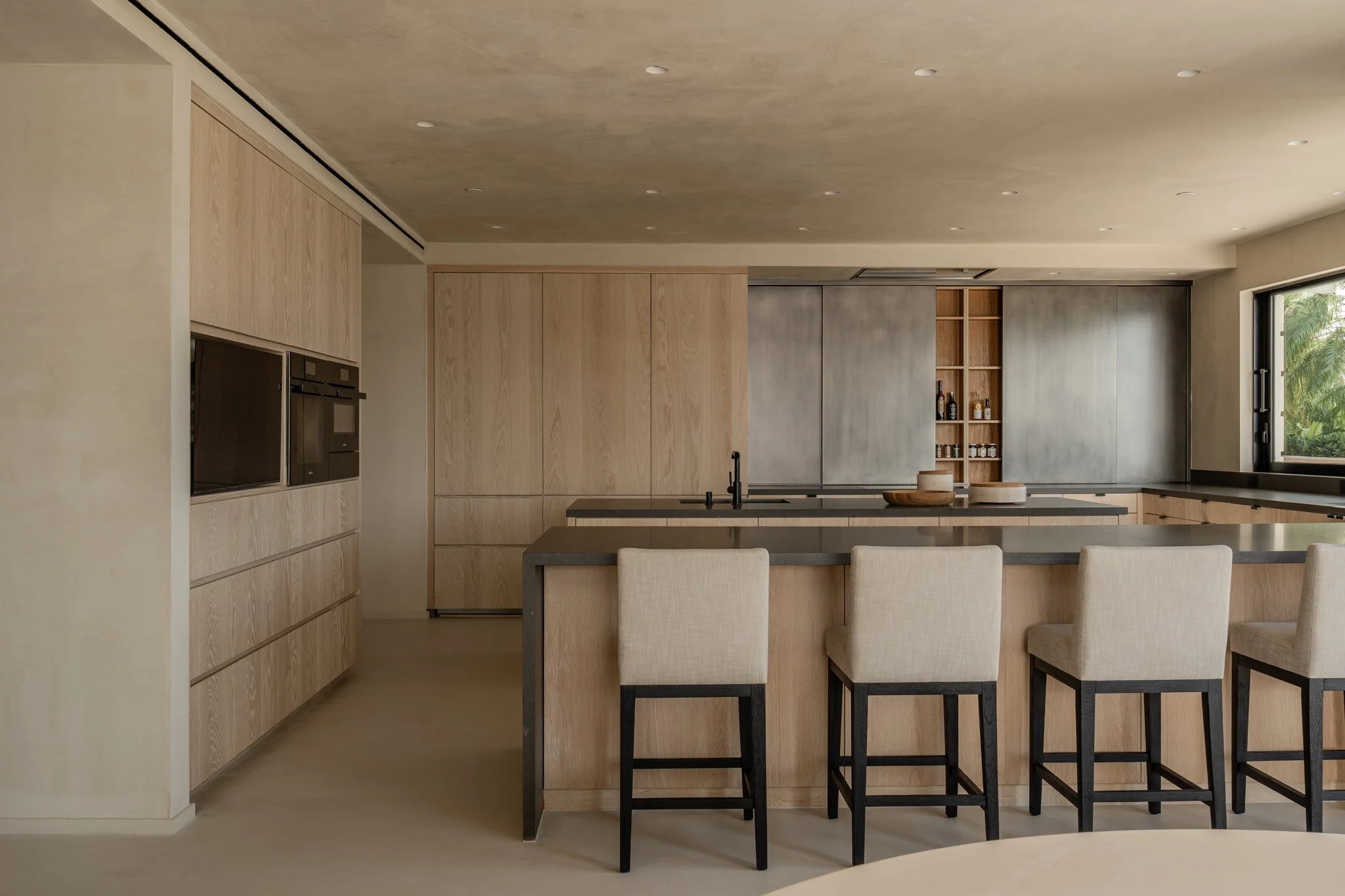 Modern kitchen with light wooden cabinets, a gray island, and beige bar stools, large window showing greenery outside.