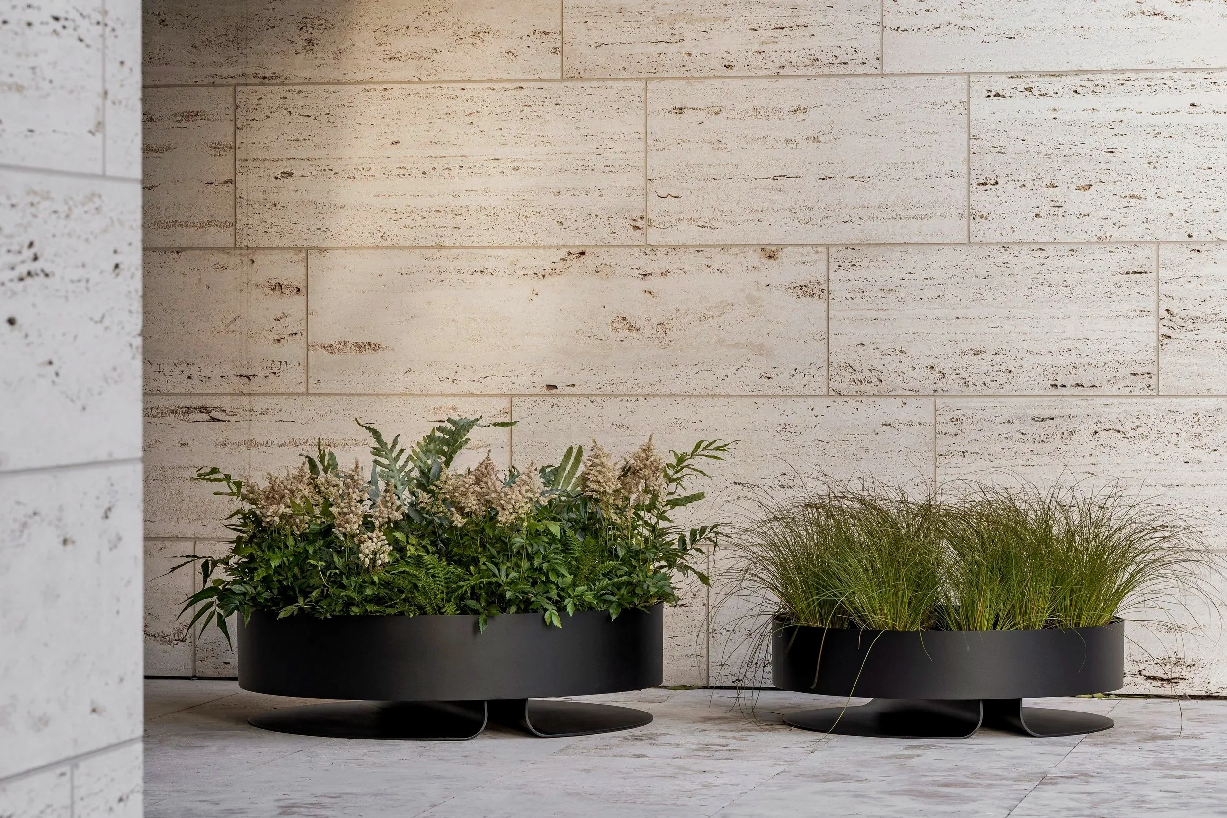 Two black planters with green plants placed on a light-colored tiled floor against a white stone wall.