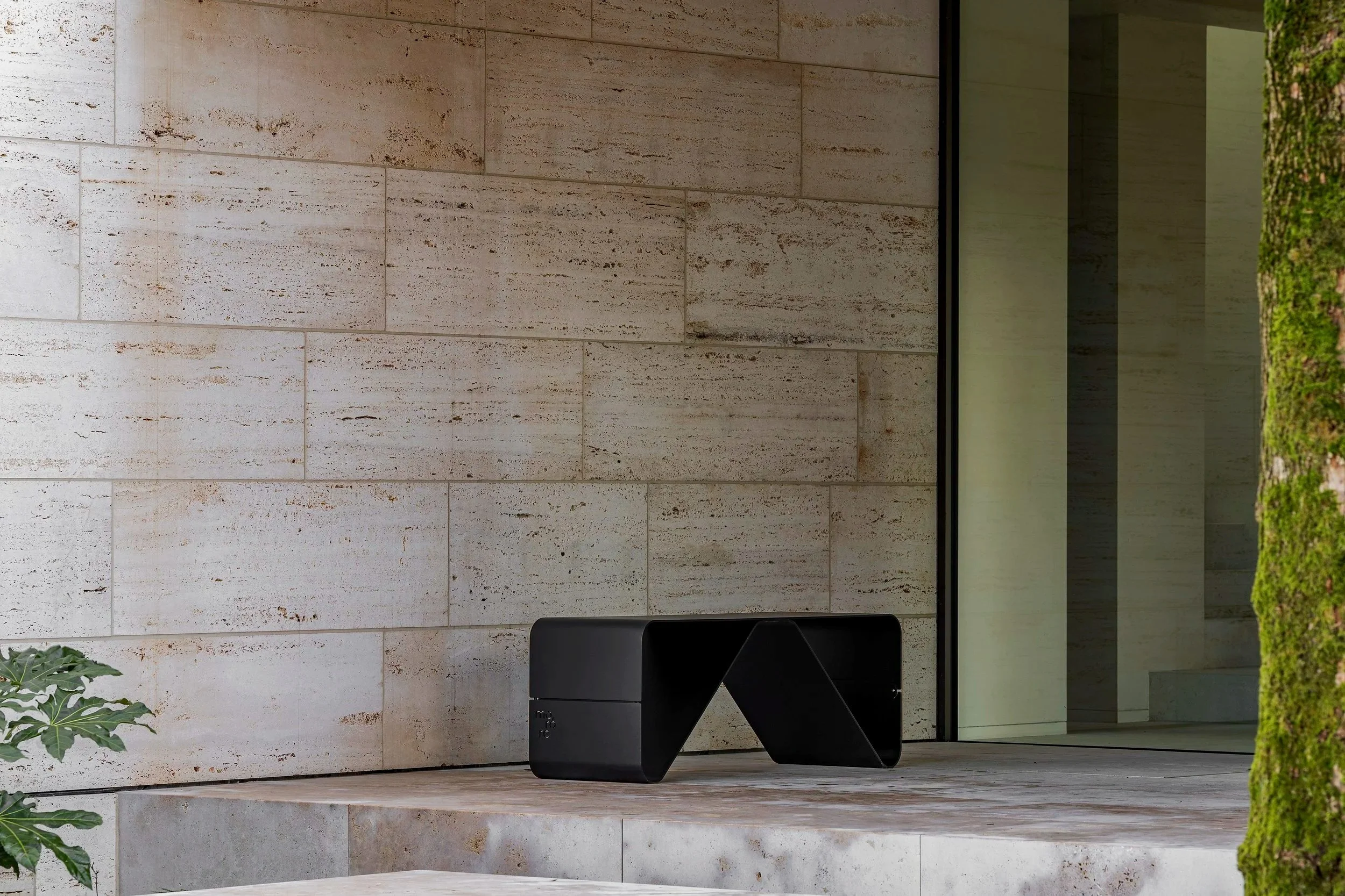 Modern black bench on concrete patio outside a building with beige stone walls, glass door, and some greenery on the side.