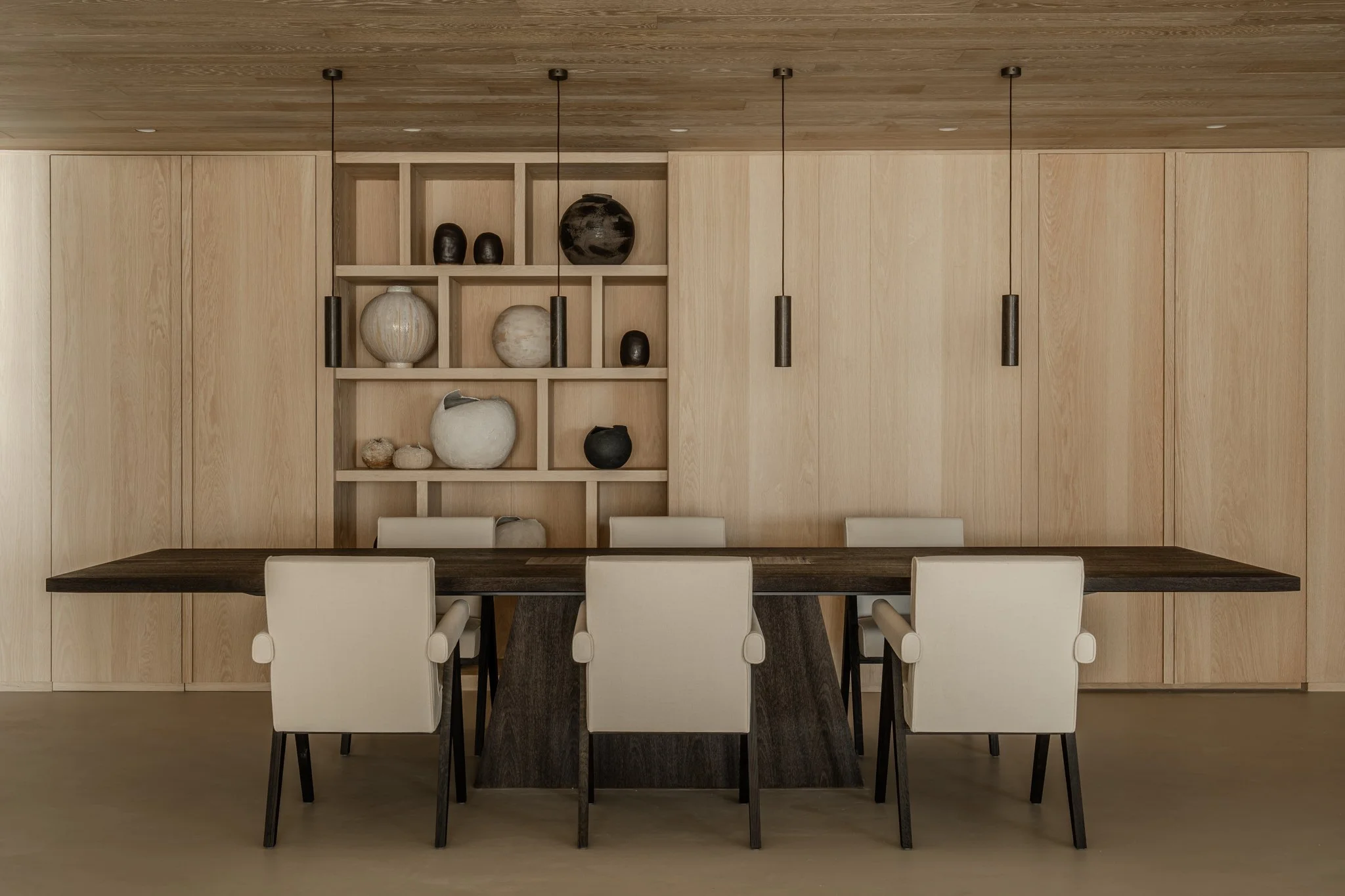 Modern dining room with light wood paneled walls and ceiling, six white upholstered chairs around a dark wood table, and a built-in wood shelf with decorative vases.
