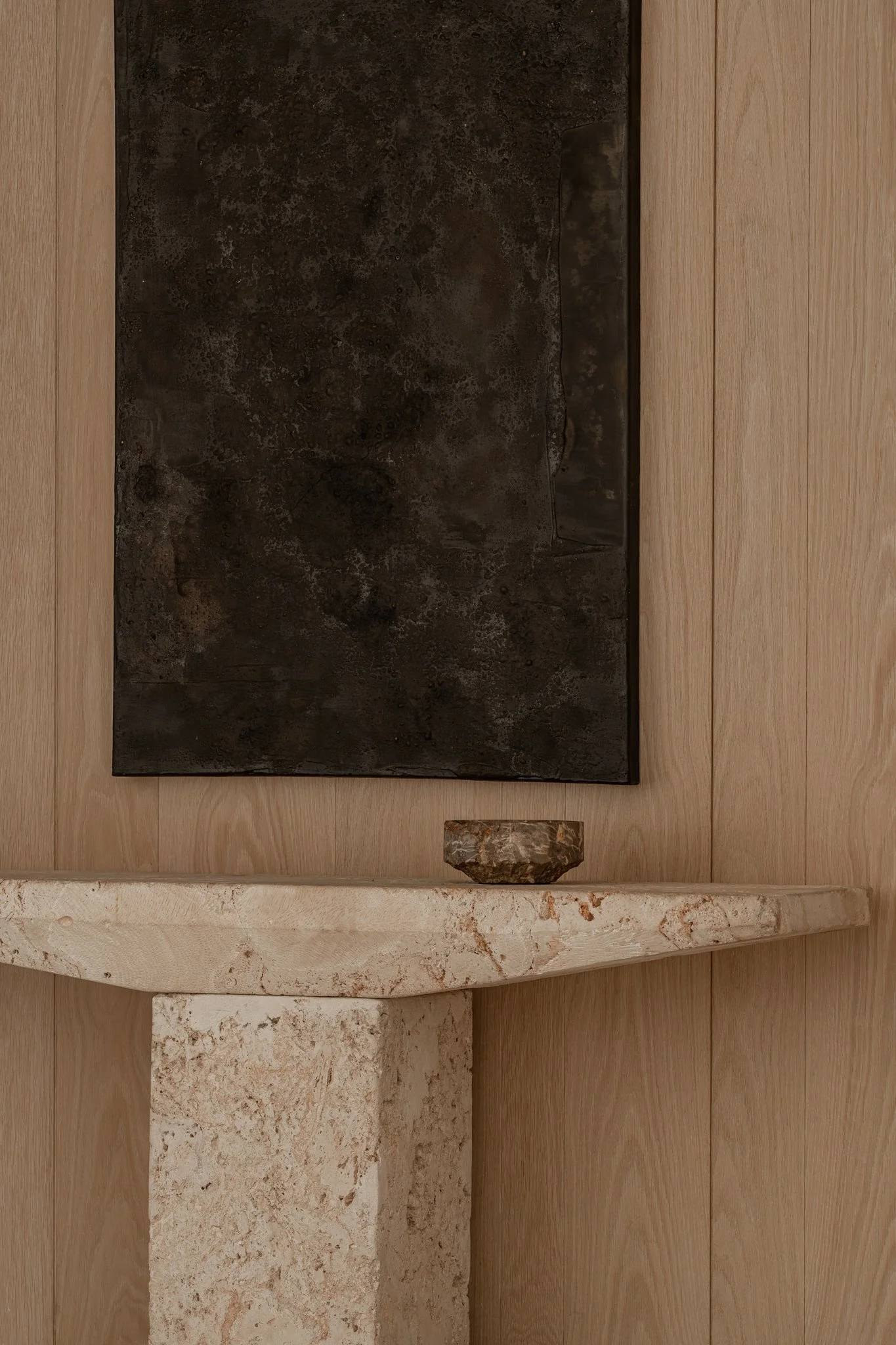 Close-up of a black, textured artwork hanging on a wooden wall above a cream-colored marble shelf with a small, irregularly shaped stone holder on top.