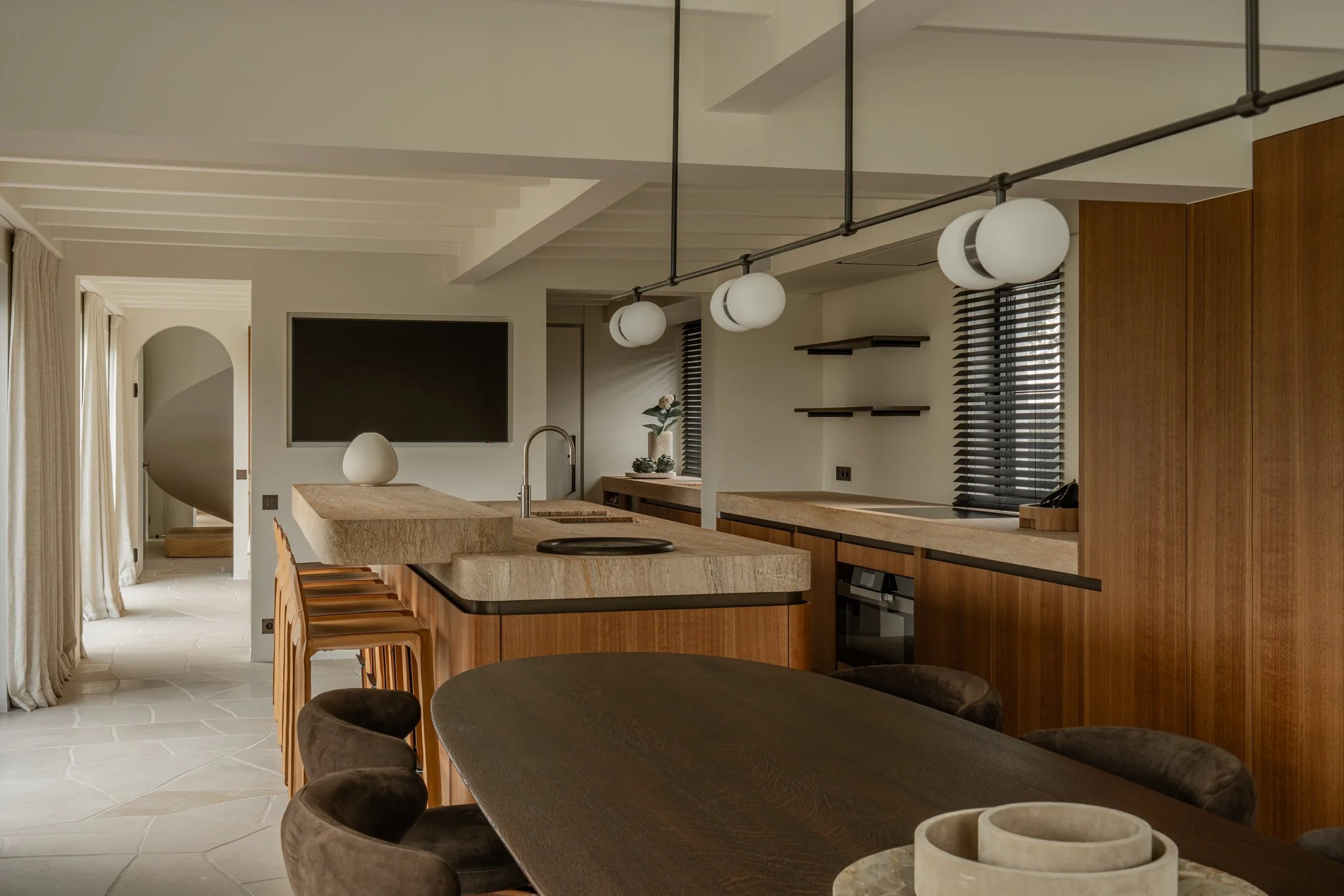 Modern kitchen with wooden cabinetry, marble countertops, round pendant lights, and a dining table with brown chairs.